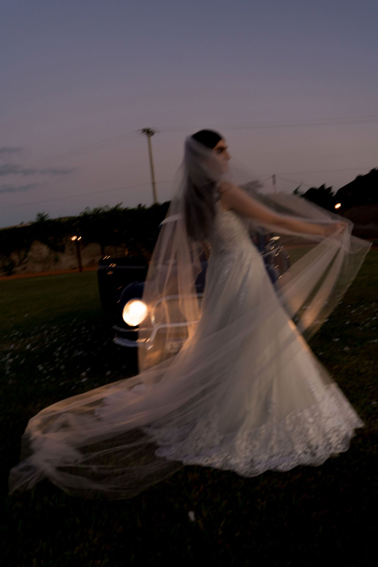 Bride in white lace gown and veil twirling outdoors at dusk near a vintage car with glowing headlight.