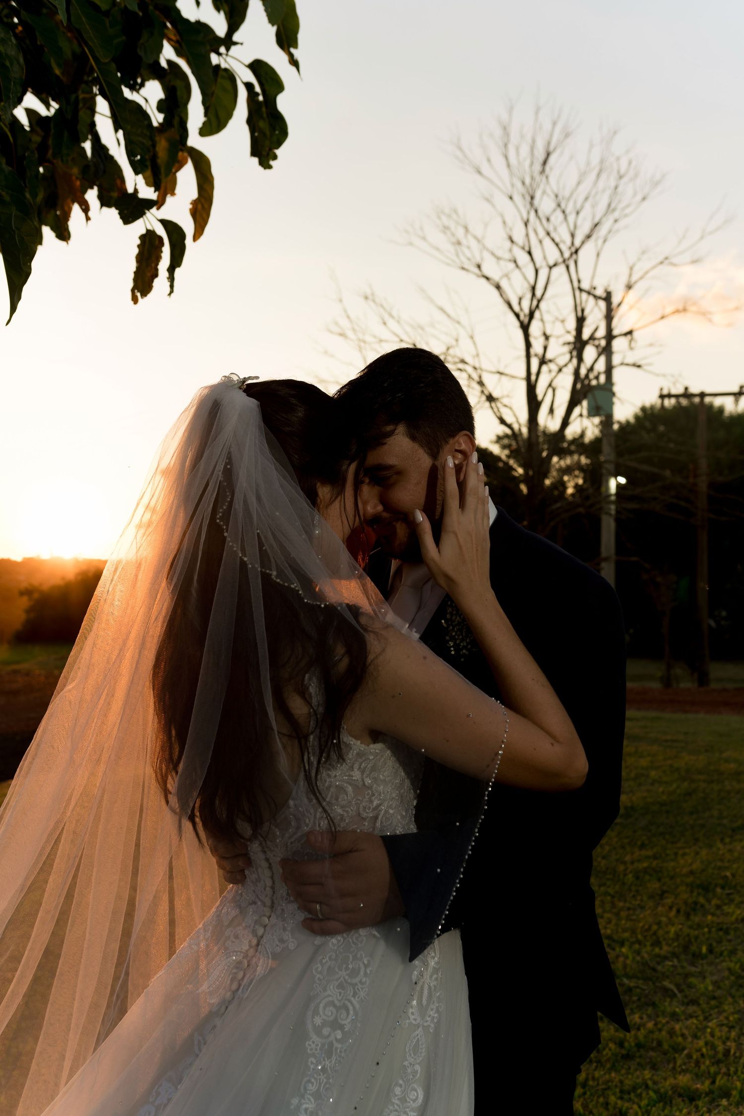 Bride and groom share a romantic sunset kiss outdoors, bride wearing lace gown and veil, groom in dark suit.