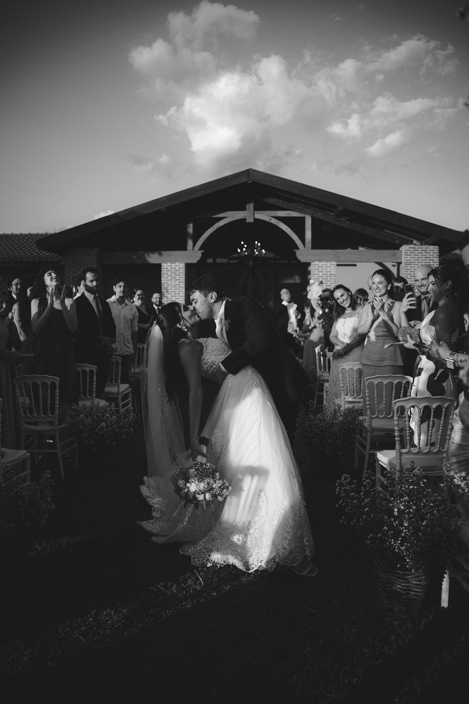 Black and white photo of a bride and groom kissing at the altar, surrounded by applauding wedding guests outdoors.
