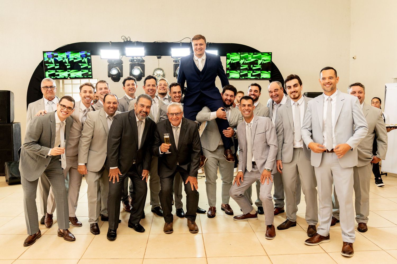 Large group of groomsmen in gray suits celebrating at a wedding reception, lifting the groom in the air.