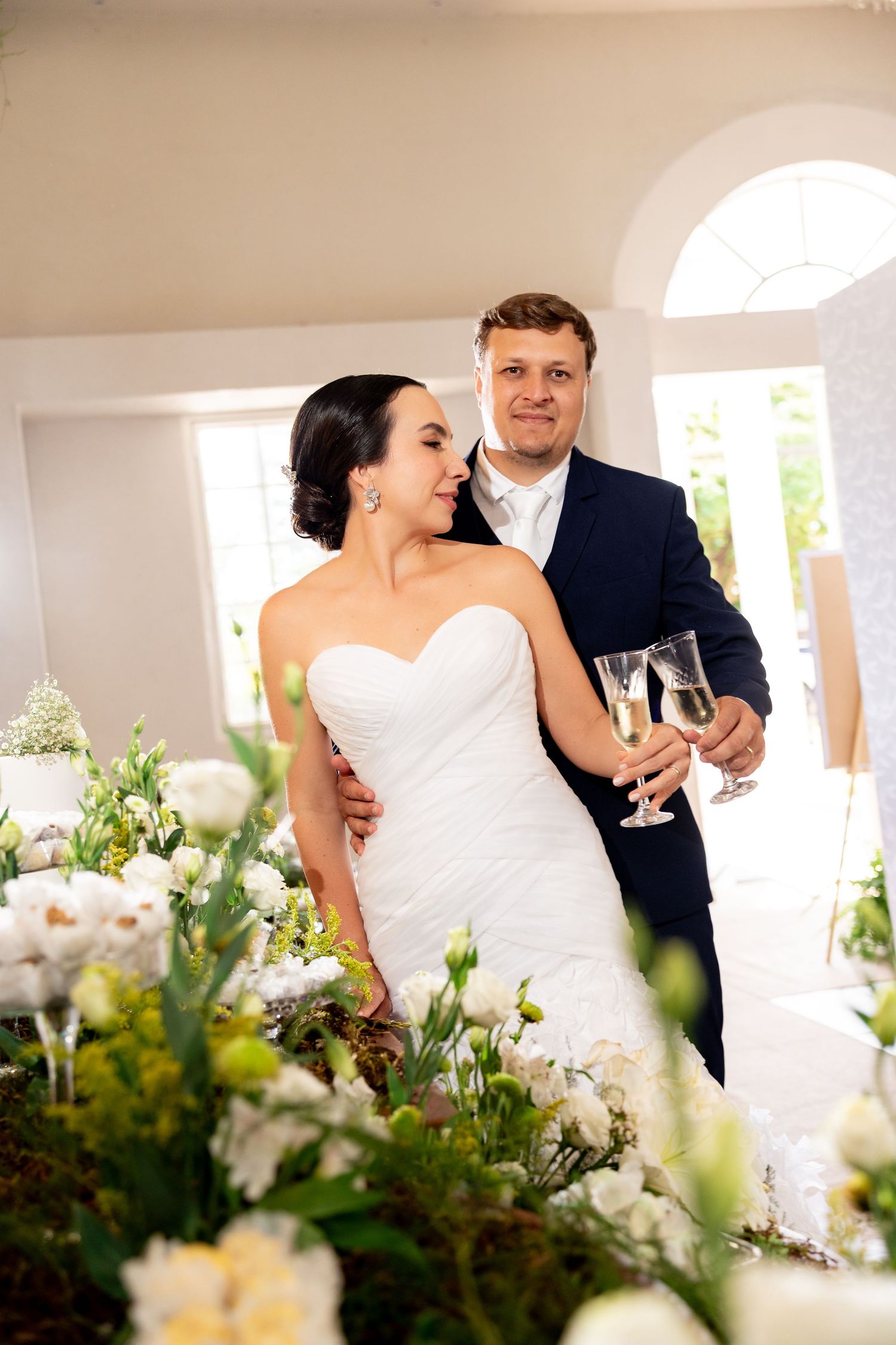 Bride and groom holding champagne glasses surrounded by white floral arrangements at their elegant wedding reception.