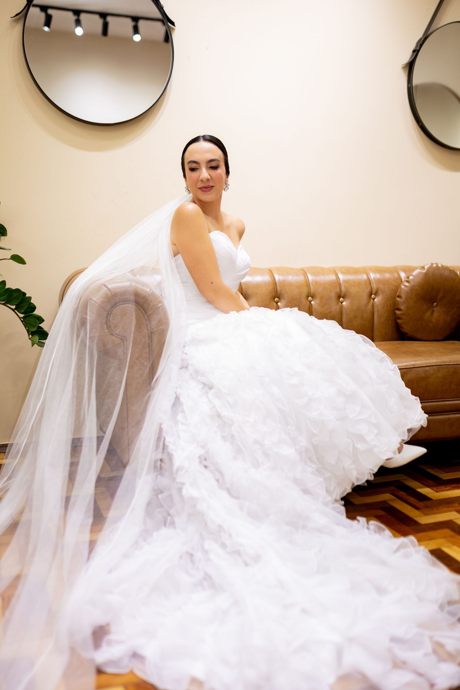 Bride in a stunning white ball gown with veil sits gracefully on a brown leather sofa in a bridal boutique.