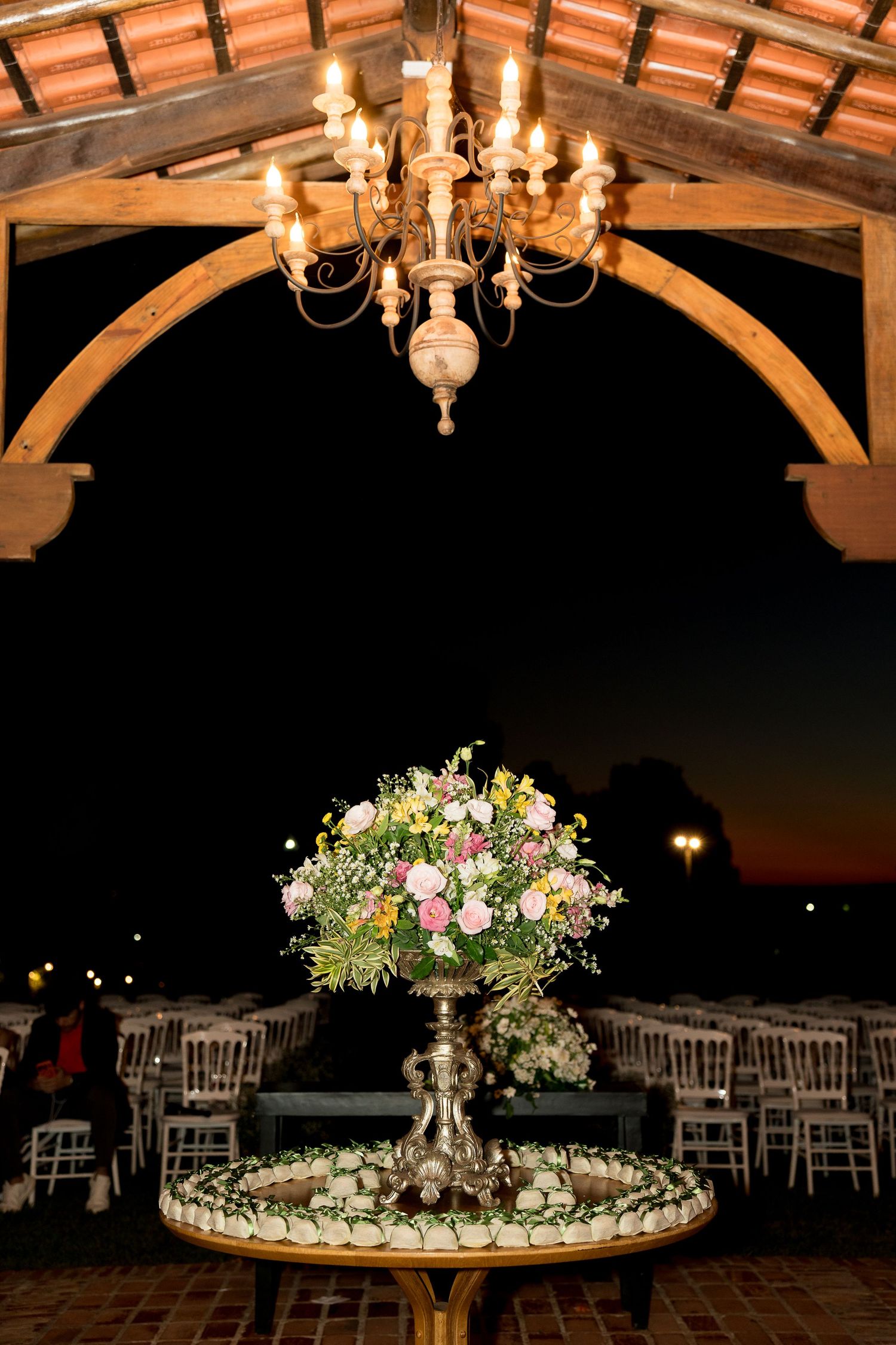 Elegant outdoor wedding venue at night with floral centerpiece, chandelier, and white chairs under a wooden pavilion.