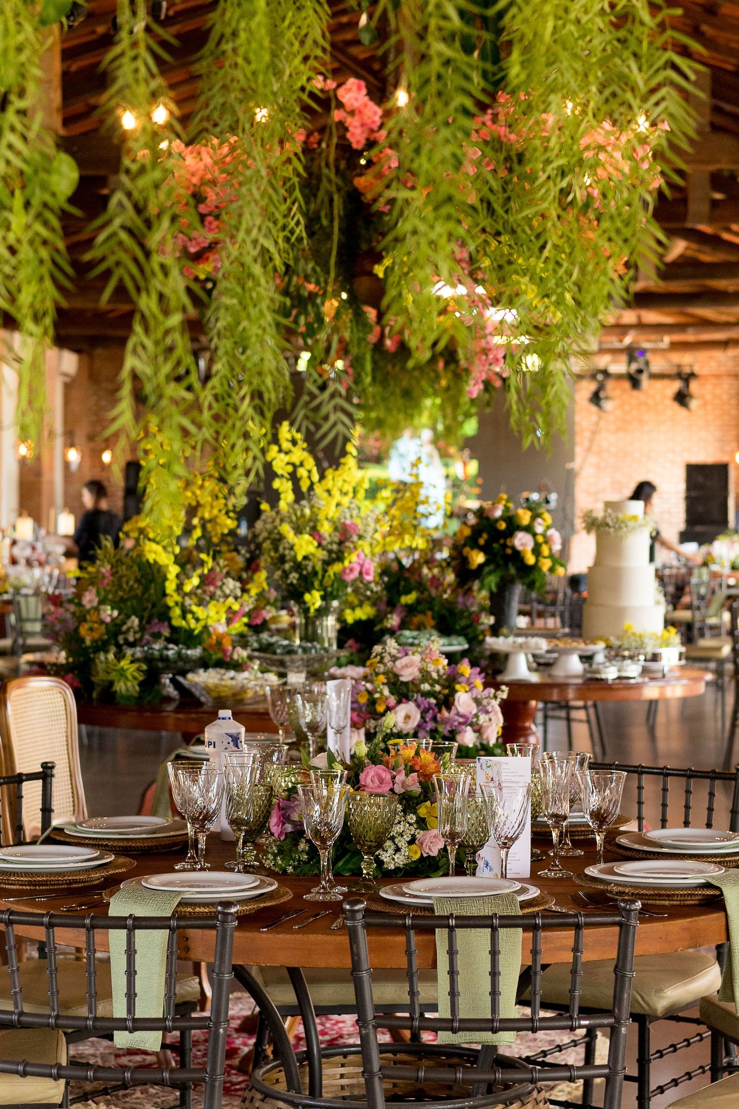 Elegant wedding reception with lush hanging greenery, floral centerpieces, crystal glassware, and a white tiered cake in a rustic venue.