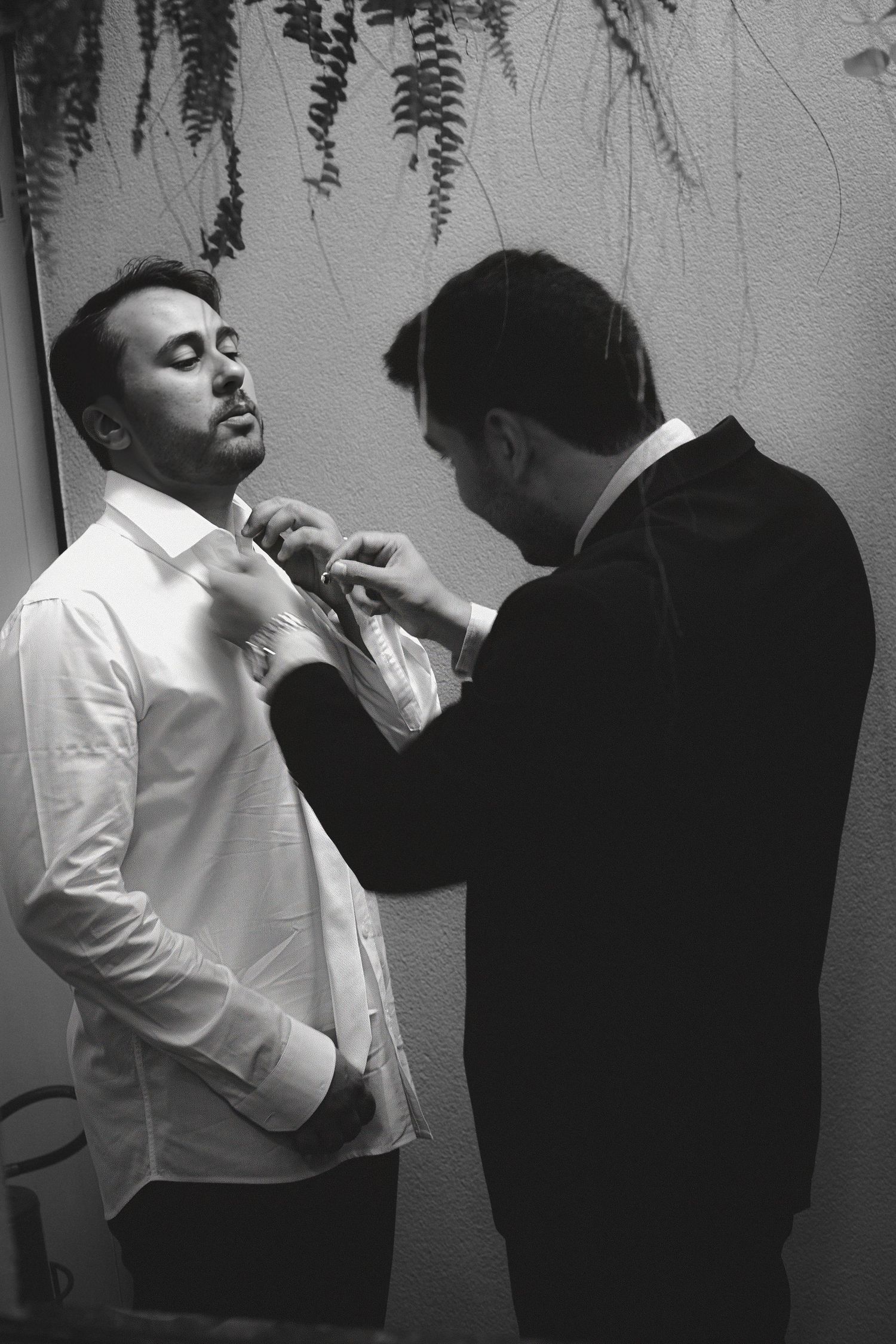 Black and white photo of a man in a suit helping another man in a white shirt with his tie before a formal event.
