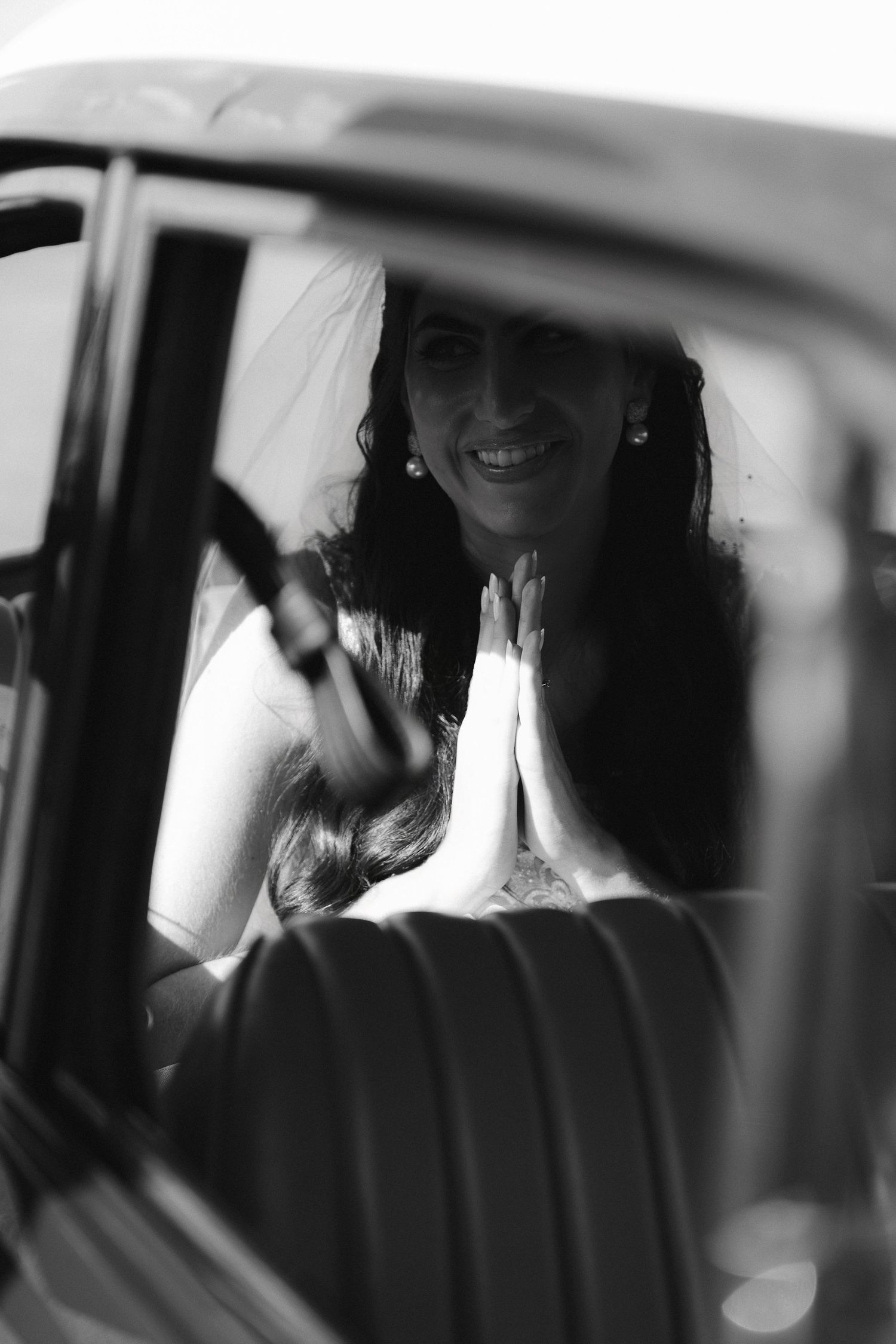 Black and white photo of a smiling bride in veil, hands pressed together in prayer pose, seen through a car window.
