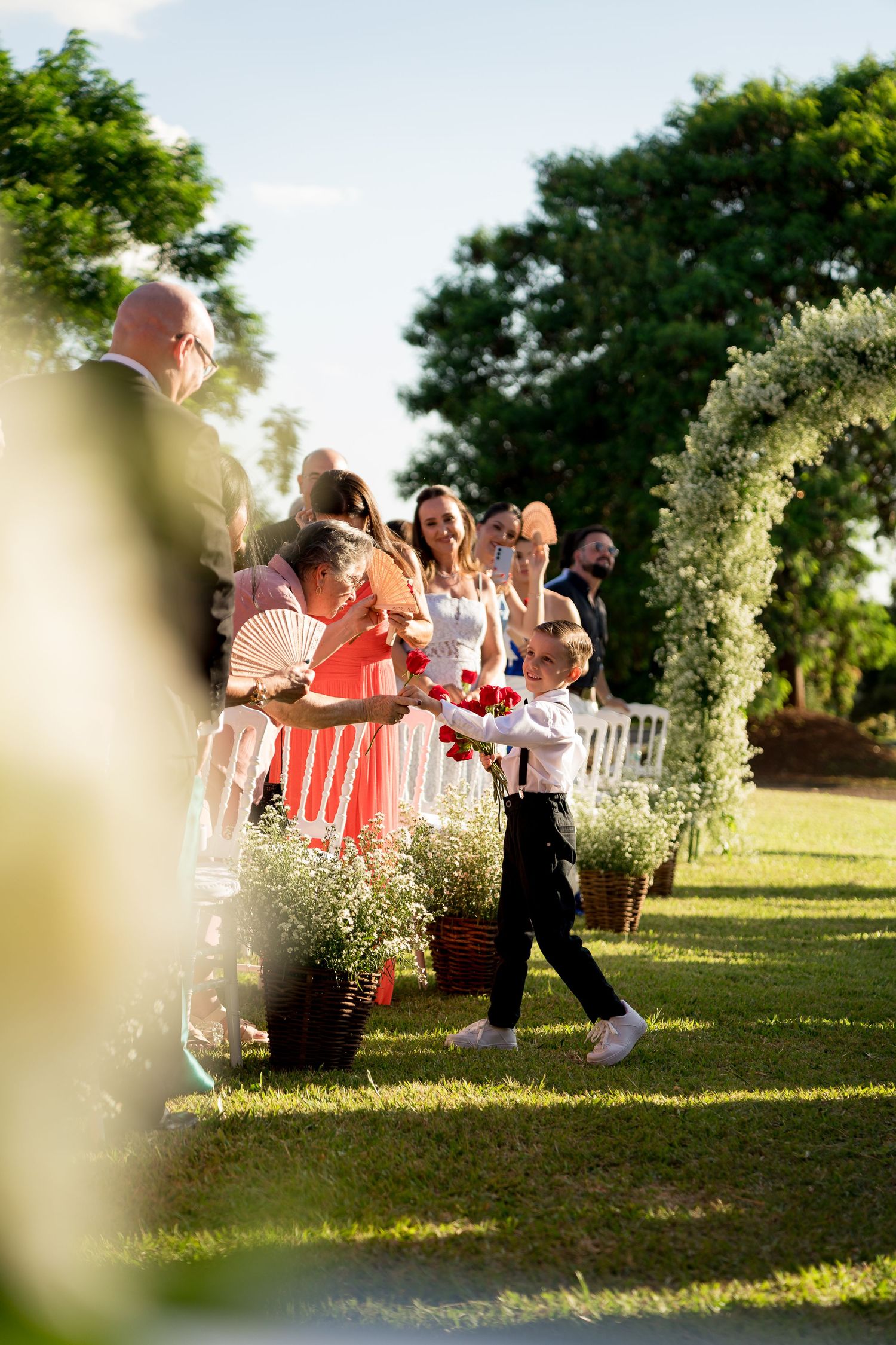 Young ring bearer walks down outdoor wedding aisle in black pants and white shirt, guests smiling alongside floral decorations.
