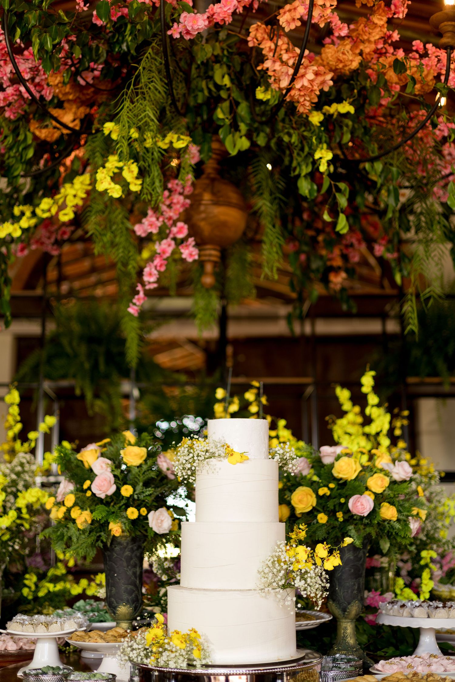 Elegant four-tier white wedding cake surrounded by yellow and pink flowers under a floral canopy.
