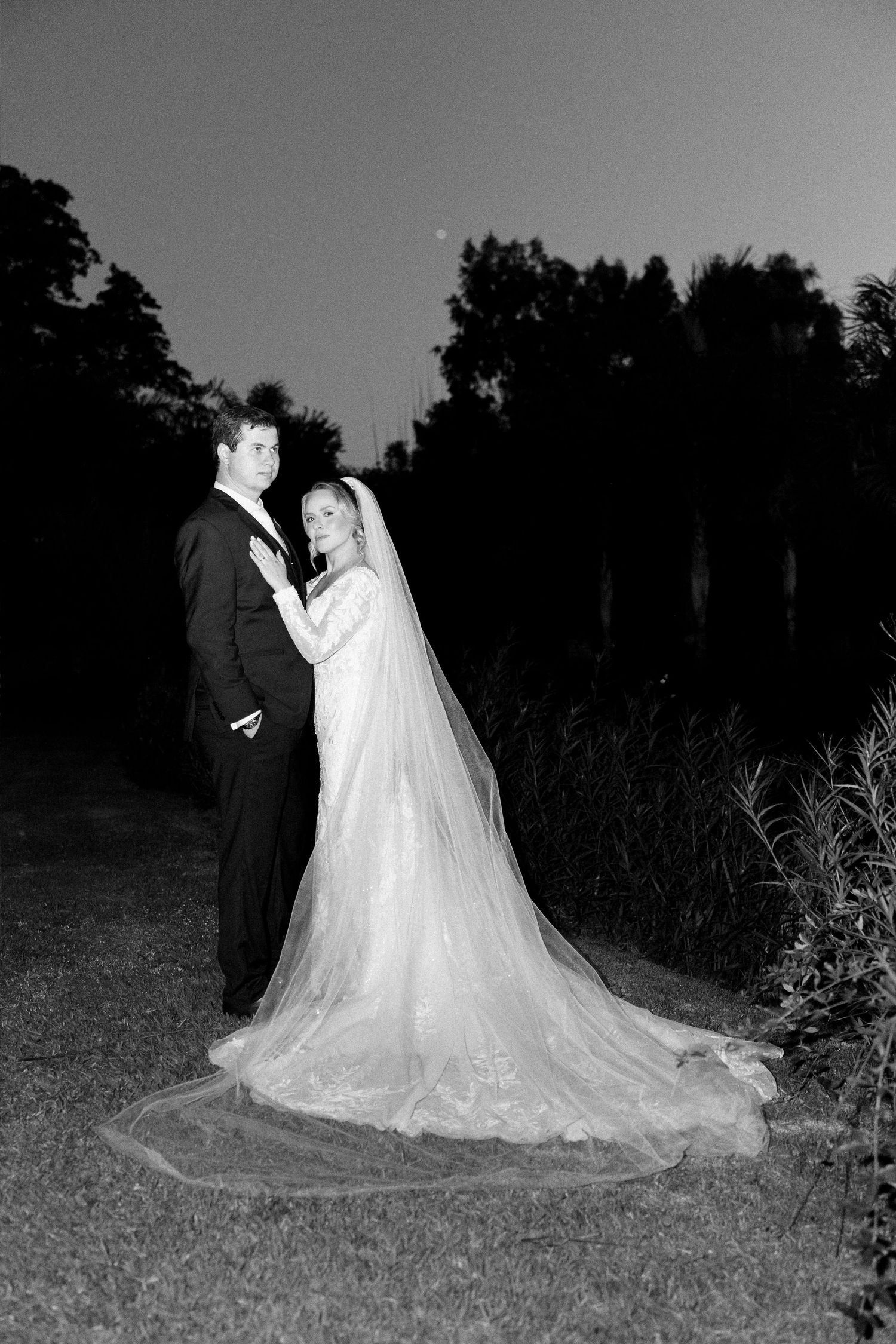 Black and white photo of a bride in a long veil and groom in a tuxedo posing outdoors at dusk.
