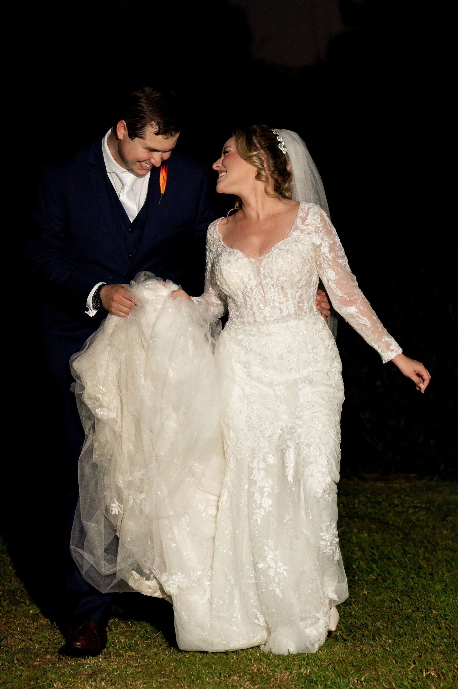 Bride and groom laughing together at night, she wears lace gown with veil, he wears navy suit with orange boutonniere.
