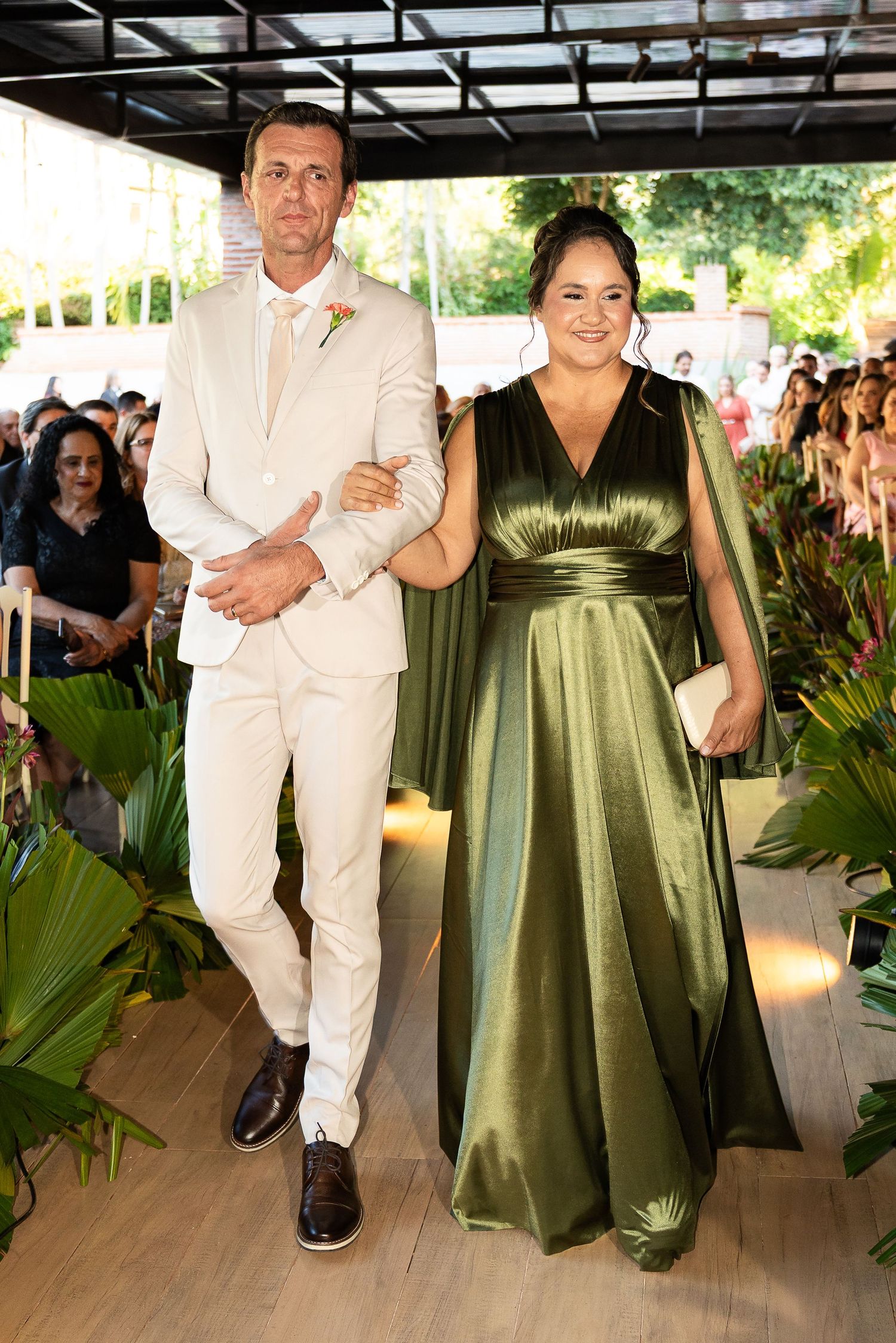 Elegant couple walking down wedding aisle, man in cream suit, woman in green satin gown, tropical decor.