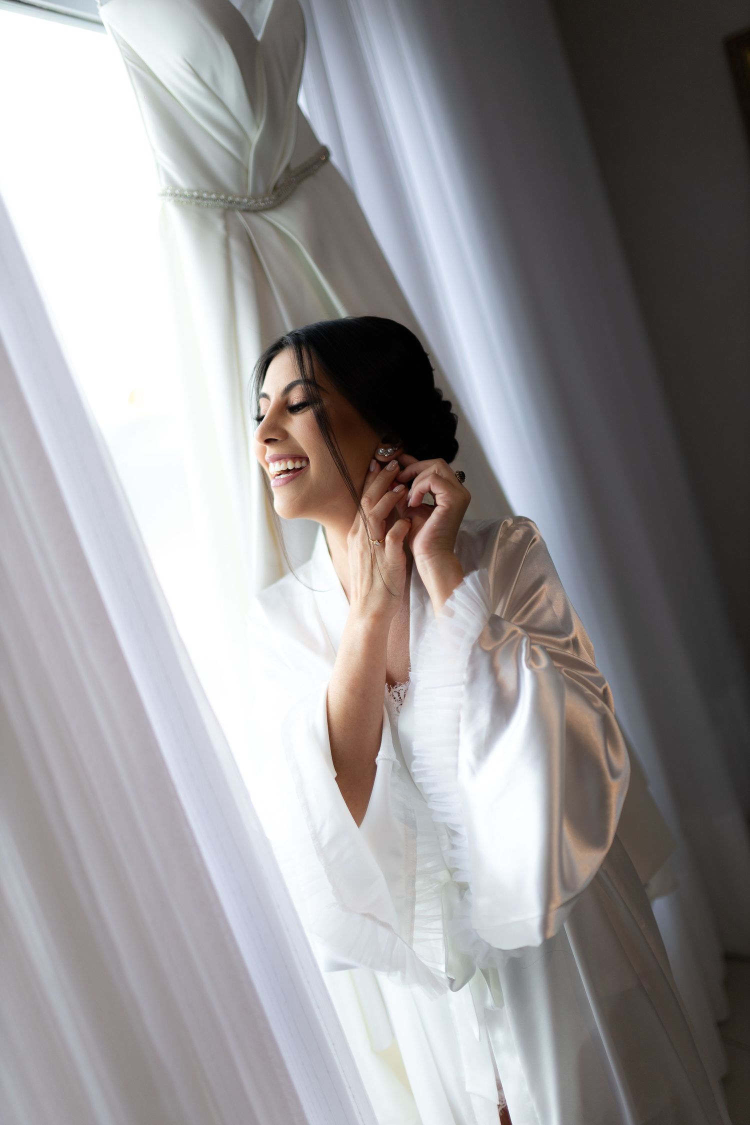 Smiling bride in white satin robe putting on earrings near window with wedding dress hanging behind.