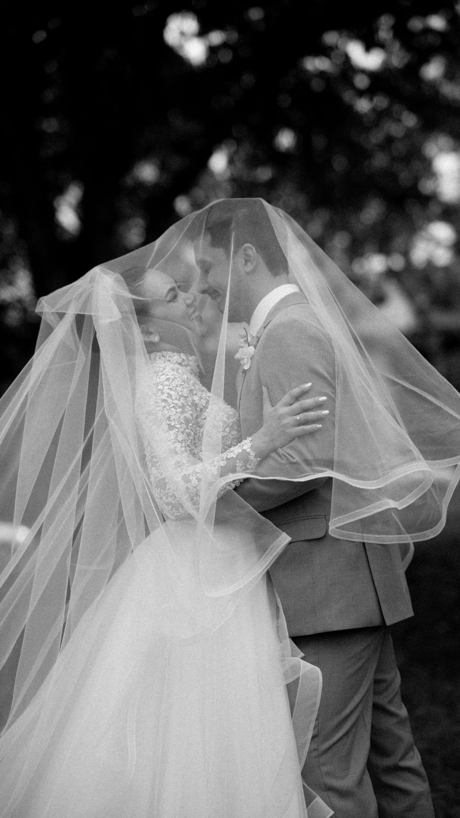 Black and white photo of bride and groom sharing a romantic kiss under a flowing wedding veil outdoors.