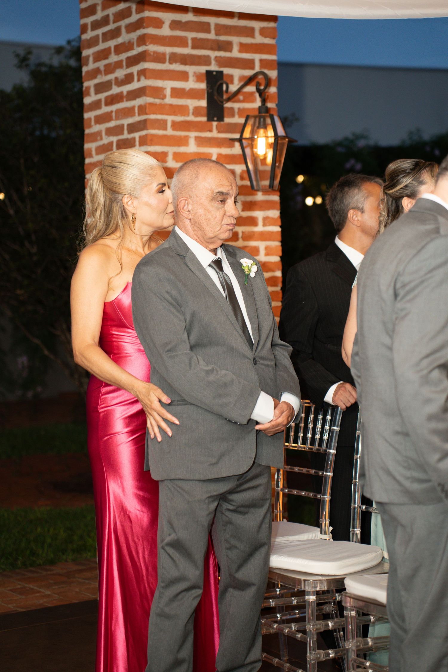 Elegant couple in red gown and gray suit at outdoor wedding reception near brick pillar at dusk.