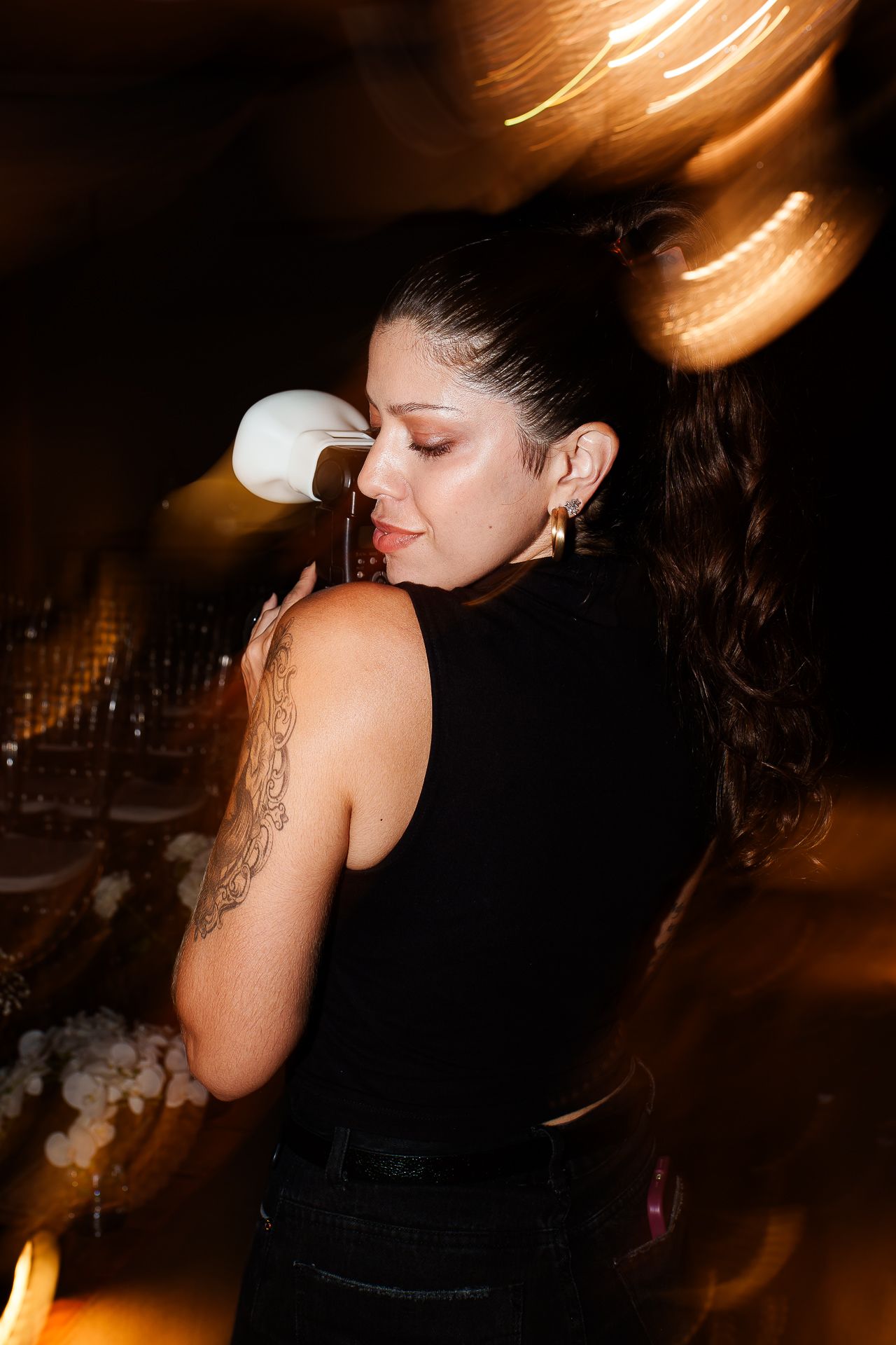 Woman with tattoo in black top holding drink at dimly lit party with bokeh lights.