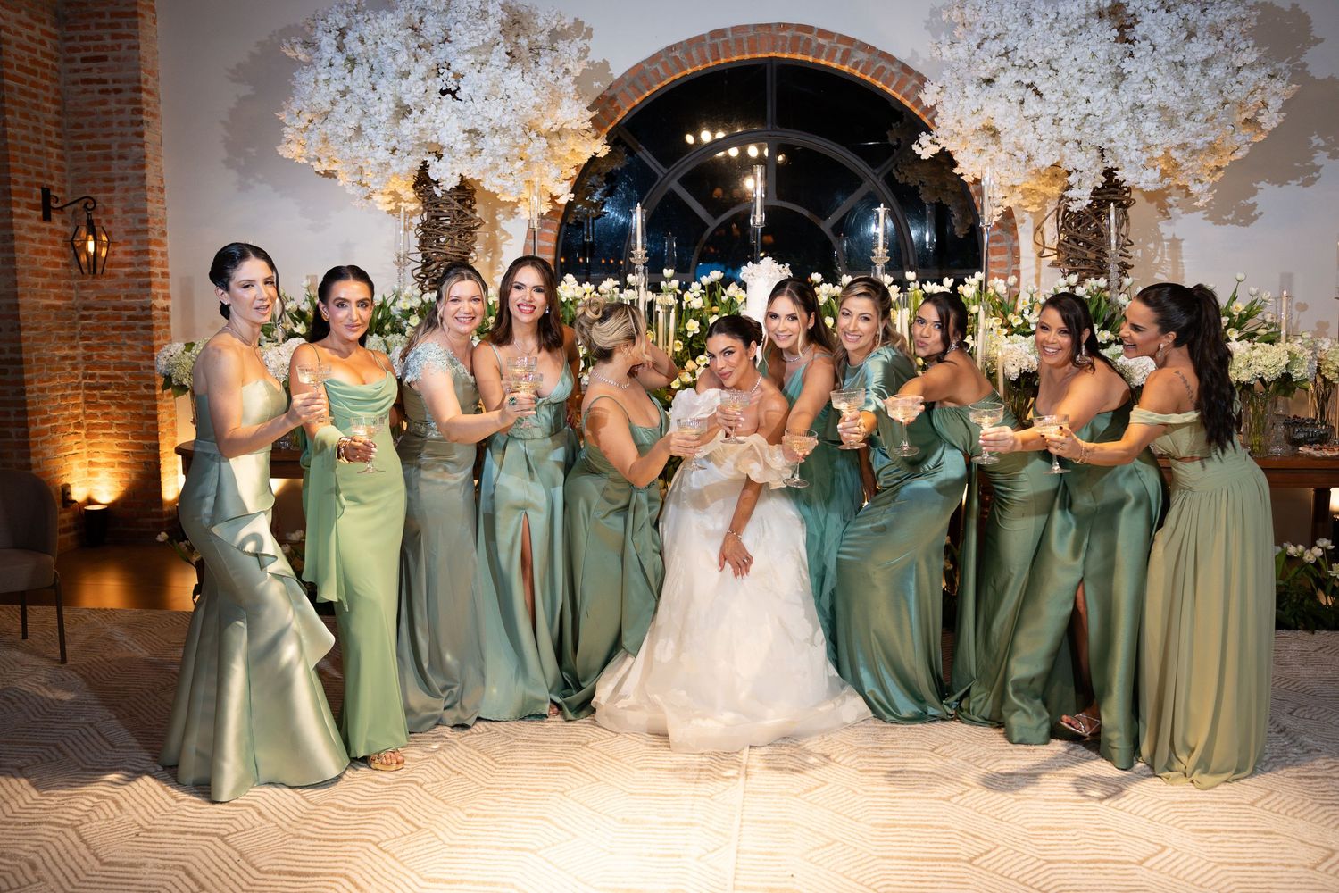 Bride in white gown poses with bridesmaids in sage green dresses at elegant wedding reception with floral arch.