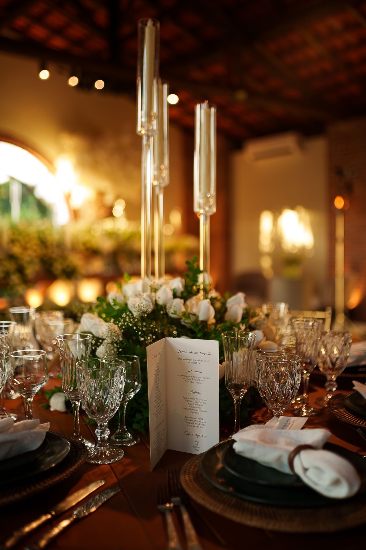 Elegant wedding reception table with crystal glasses, white roses, tall candles, and a menu card in warm candlelight.