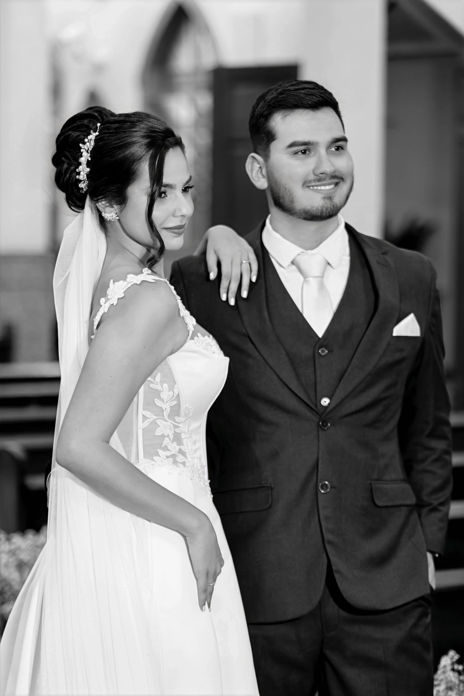 Black and white wedding photo of a smiling bride in lace gown and groom in dark suit posing together outside a church.