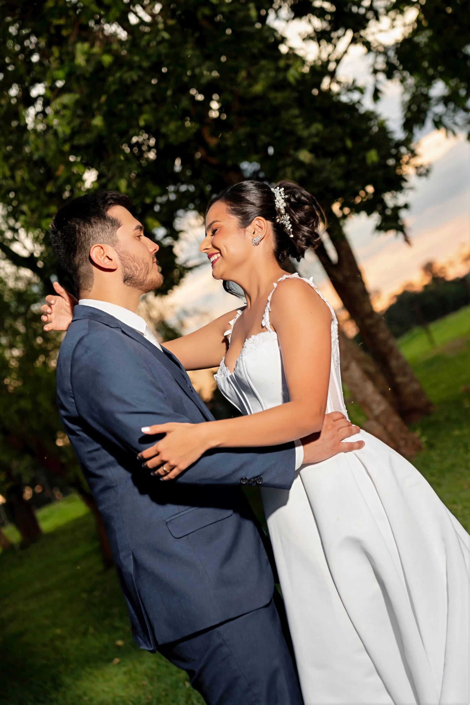 A joyful bride and groom share a romantic embrace outdoors at sunset, surrounded by lush green trees.