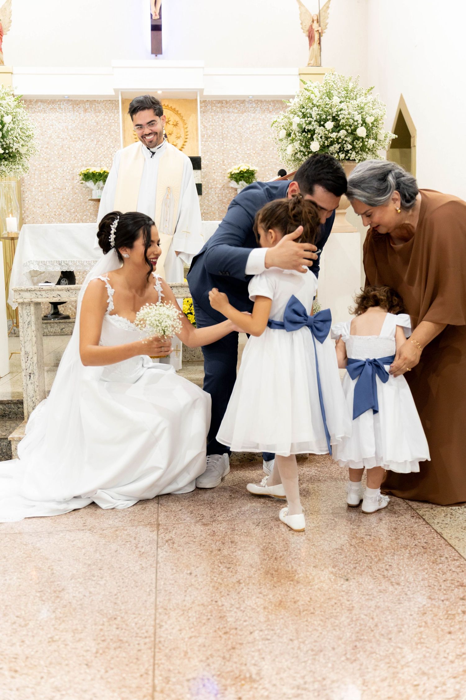 Bride and groom hugging flower girls in white dresses with blue bows during a church wedding ceremony.