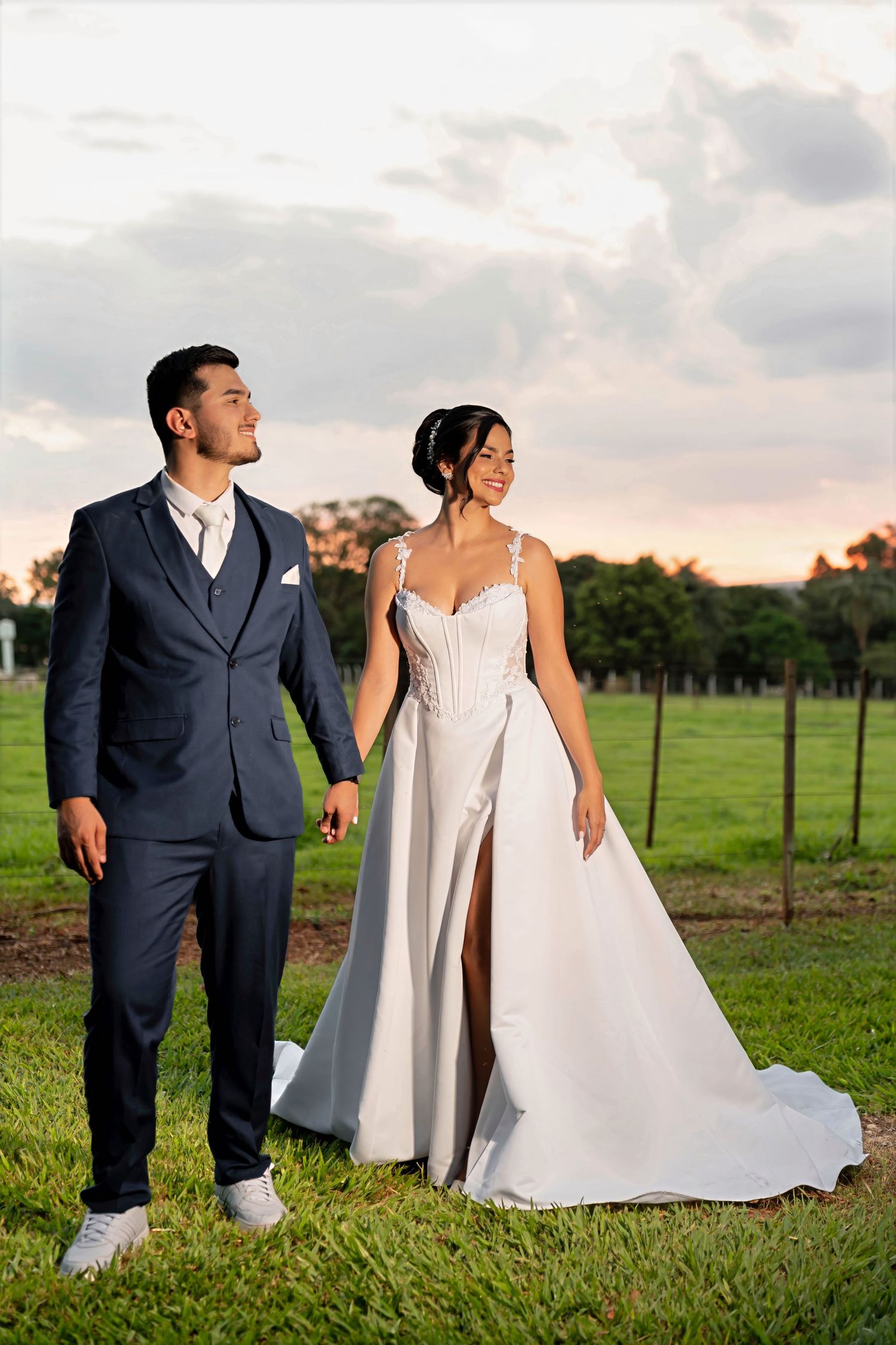 A bride and groom hold hands in a green field at sunset, she in a white gown, he in a navy suit.