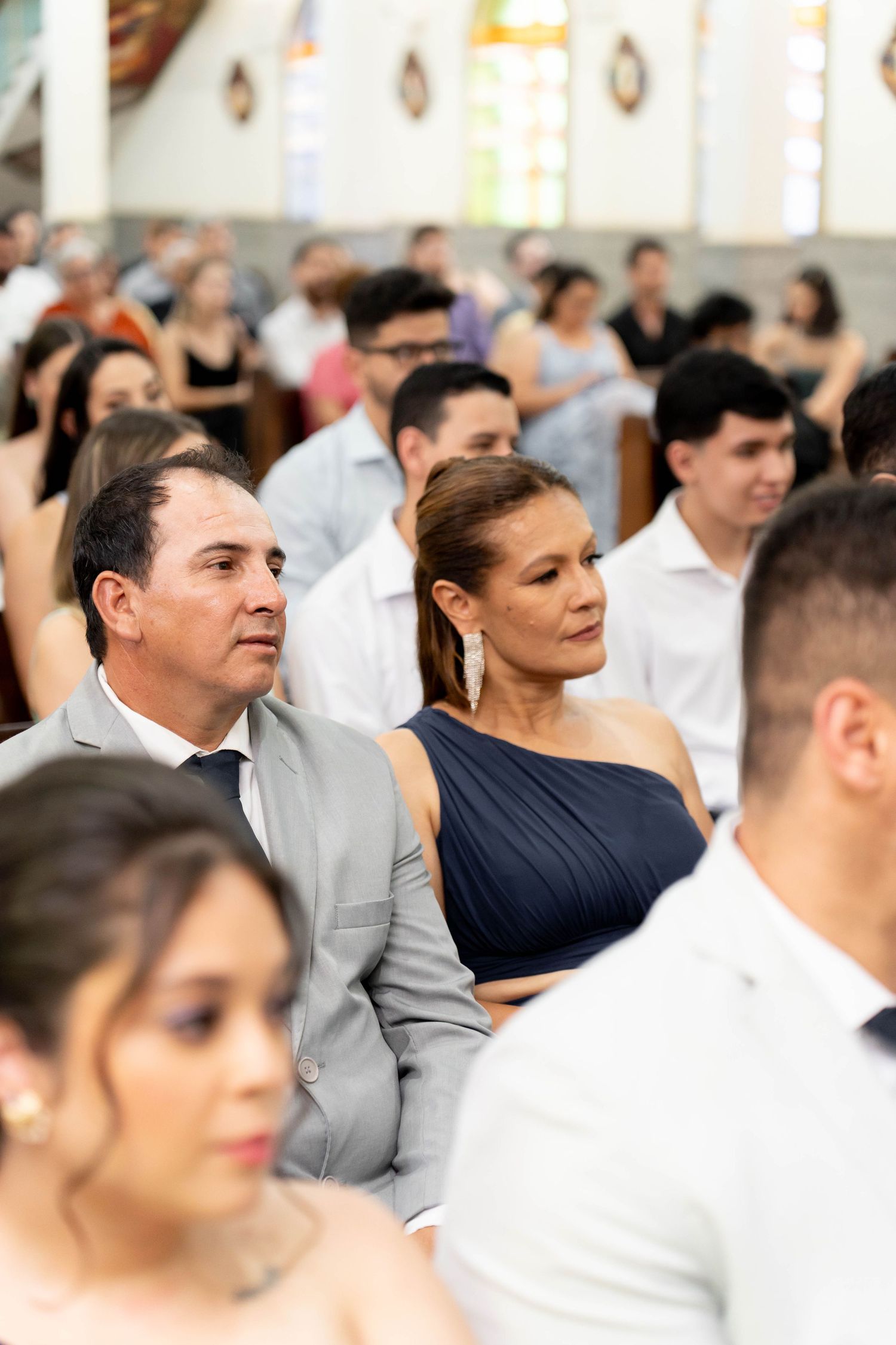 Elegantly dressed wedding guests seated in a church, including a man in gray suit and woman in navy dress.