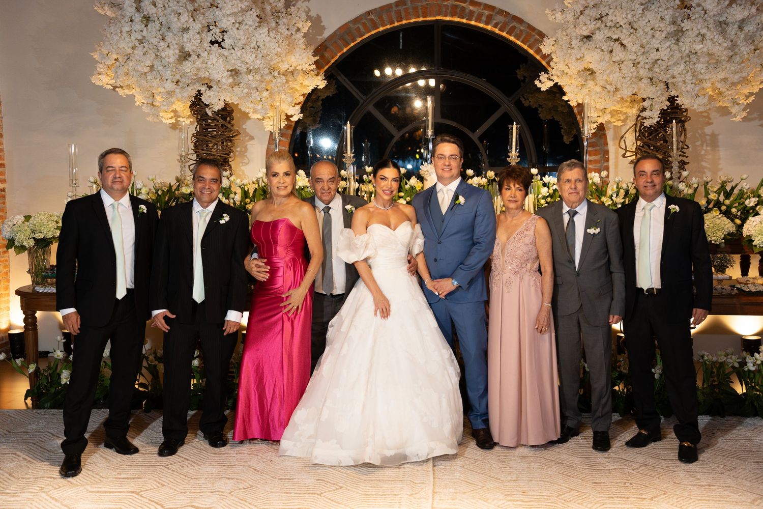 Wedding party poses together in elegant venue with floral decor, bride in white gown and groom in blue suit centered.