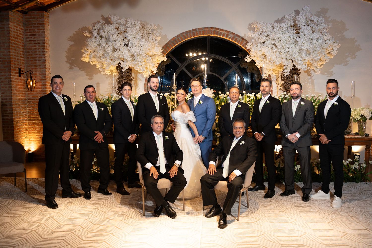 Wedding party poses with bride and groom before floral arch, groomsmen in suits flanking couple at elegant venue.