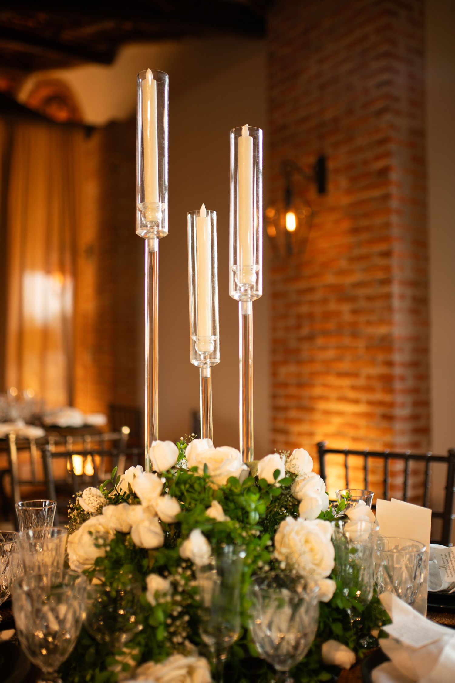 Elegant wedding reception centerpiece with white roses, greenery, and tall glass candle holders on a decorated dining table.