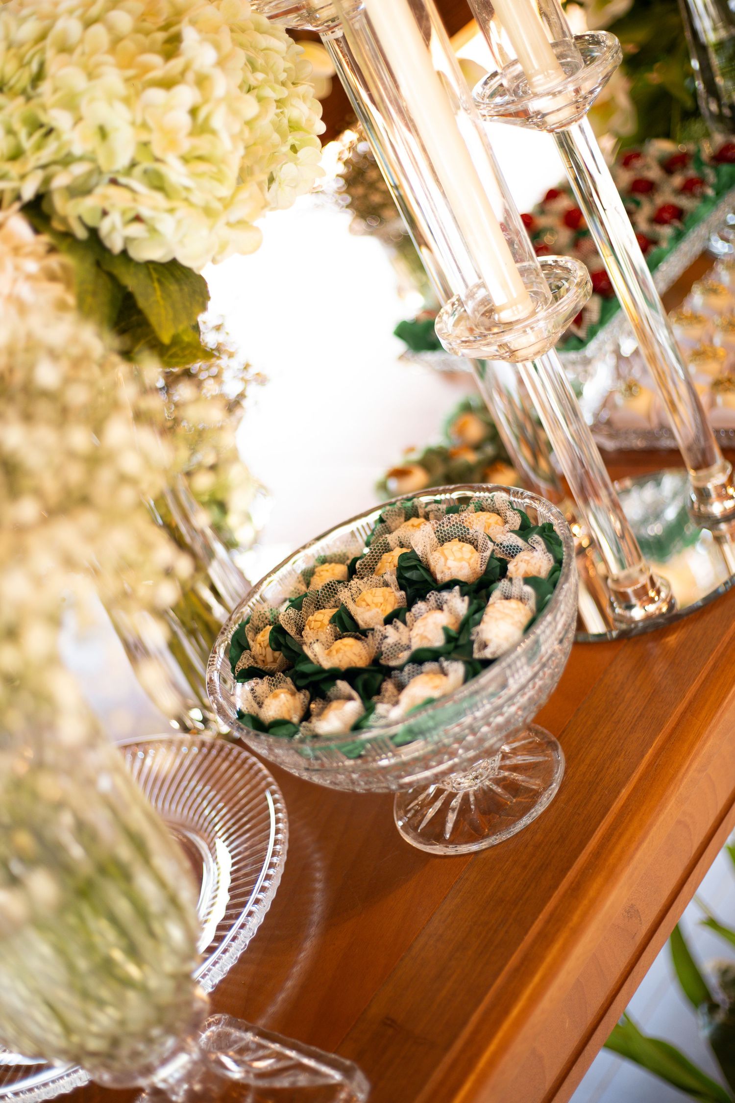 Elegant crystal dessert bowl filled with petit fours on a wooden table, surrounded by glass candelabras and white hydrangeas.
