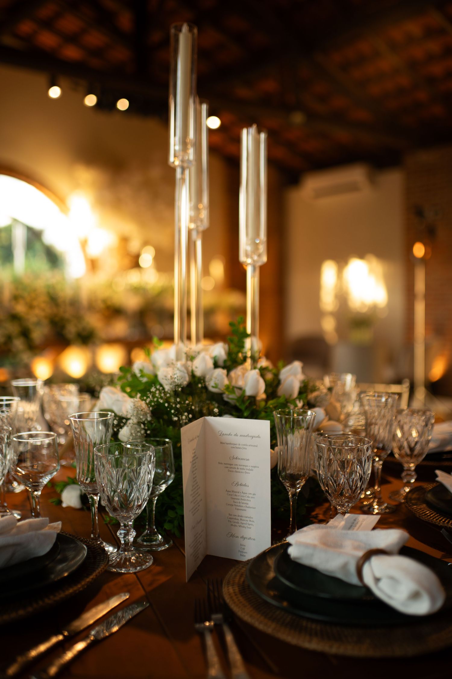 Elegant wedding reception table with crystal glasses, white roses, tall candles, and a menu card in warm candlelit ambiance.