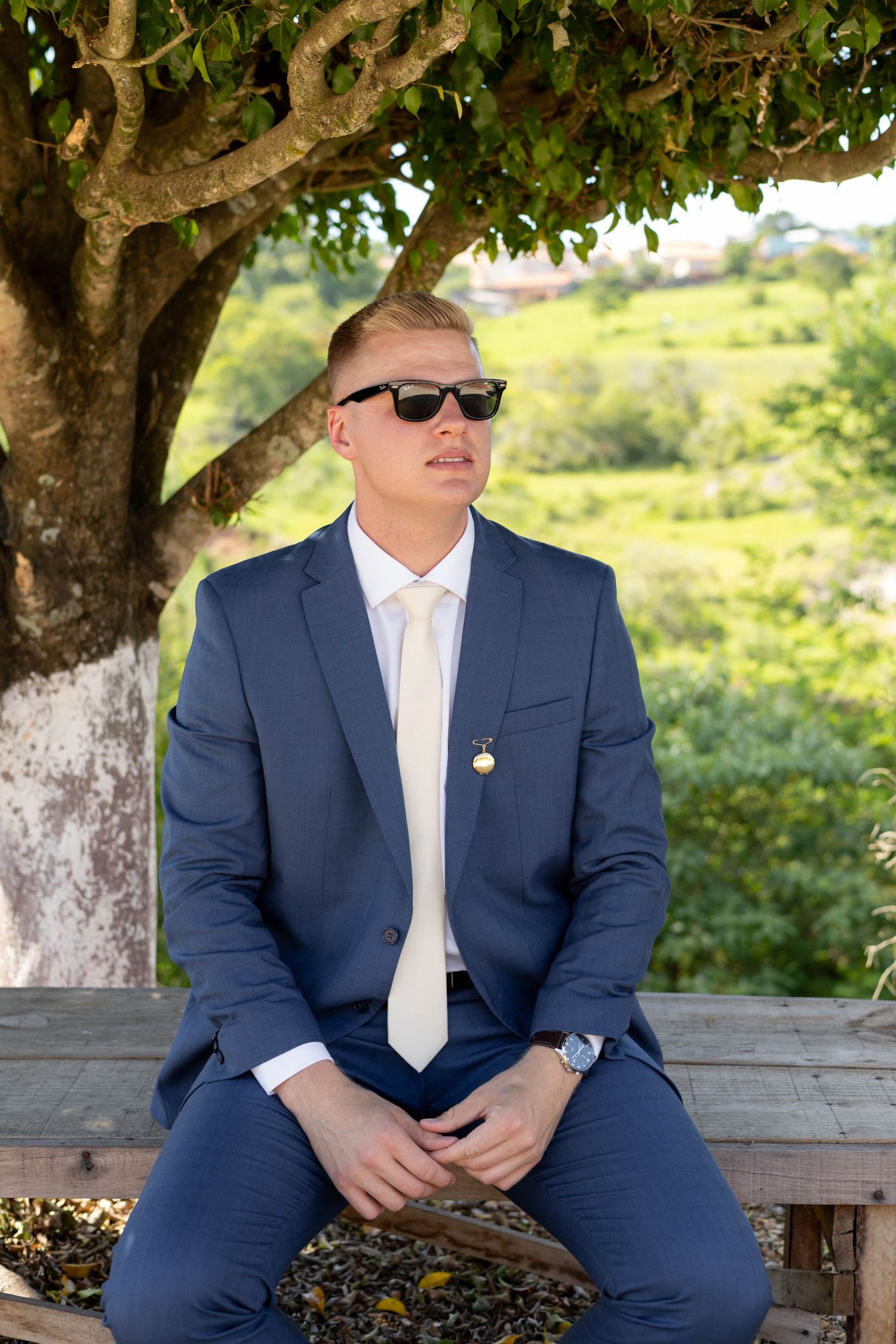 Young man in blue suit, white tie, and sunglasses sitting on wooden bench under a tree outdoors.