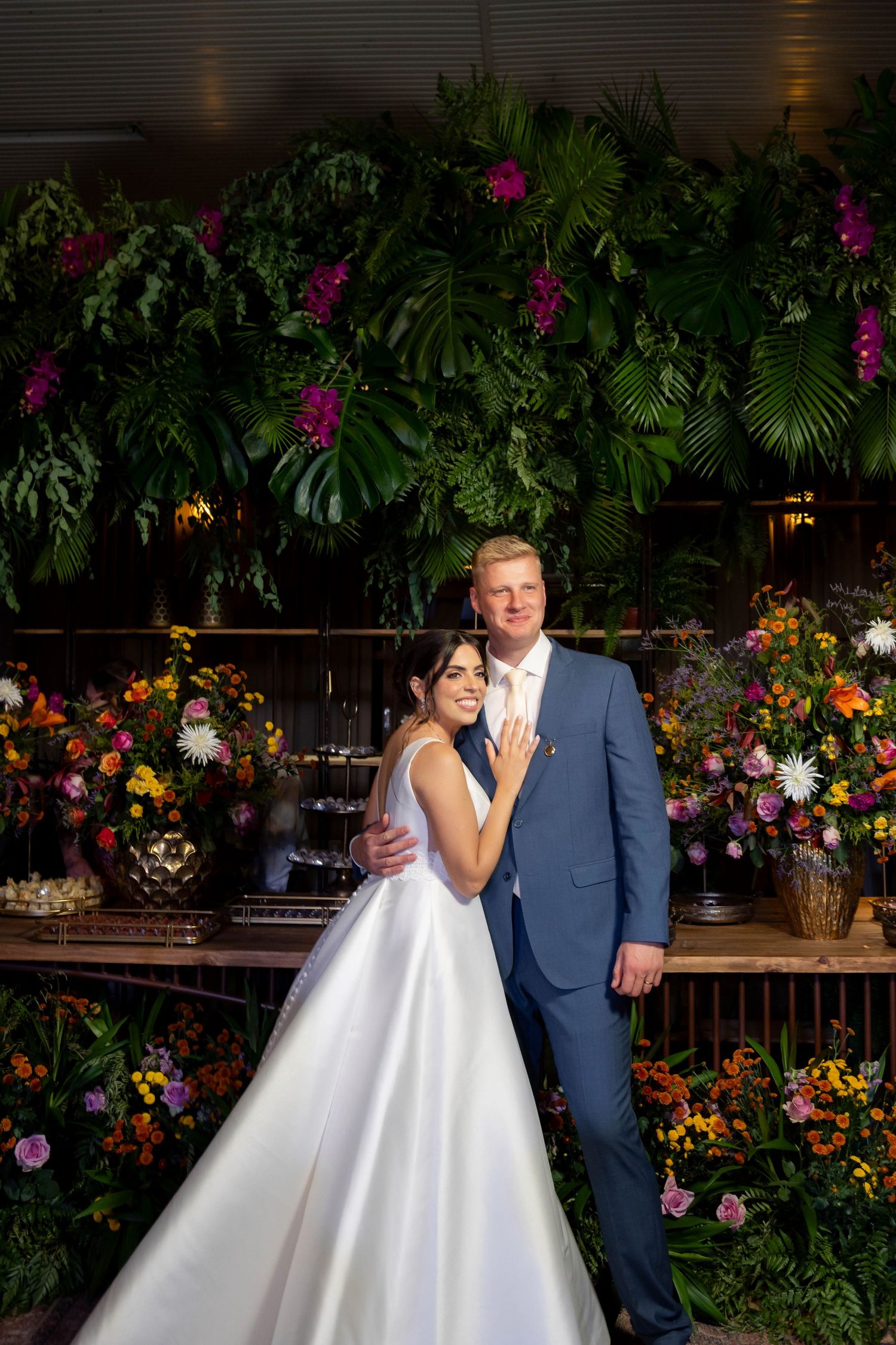 Bride and groom pose together surrounded by lush tropical greenery and colorful floral arrangements at their wedding reception.