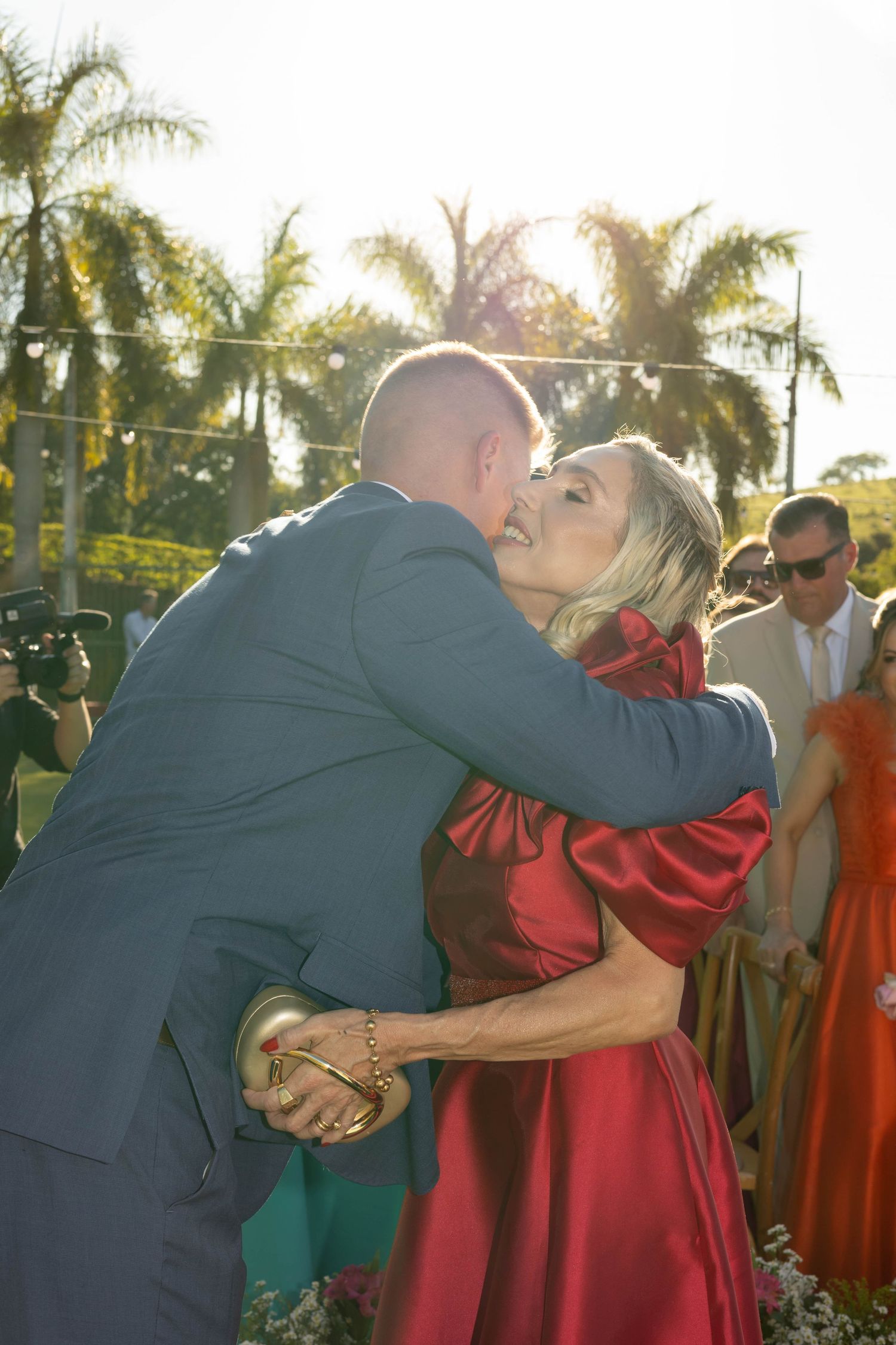 Couple sharing a kiss at an outdoor wedding, woman in red satin dress, man in gray suit, palm trees in background.