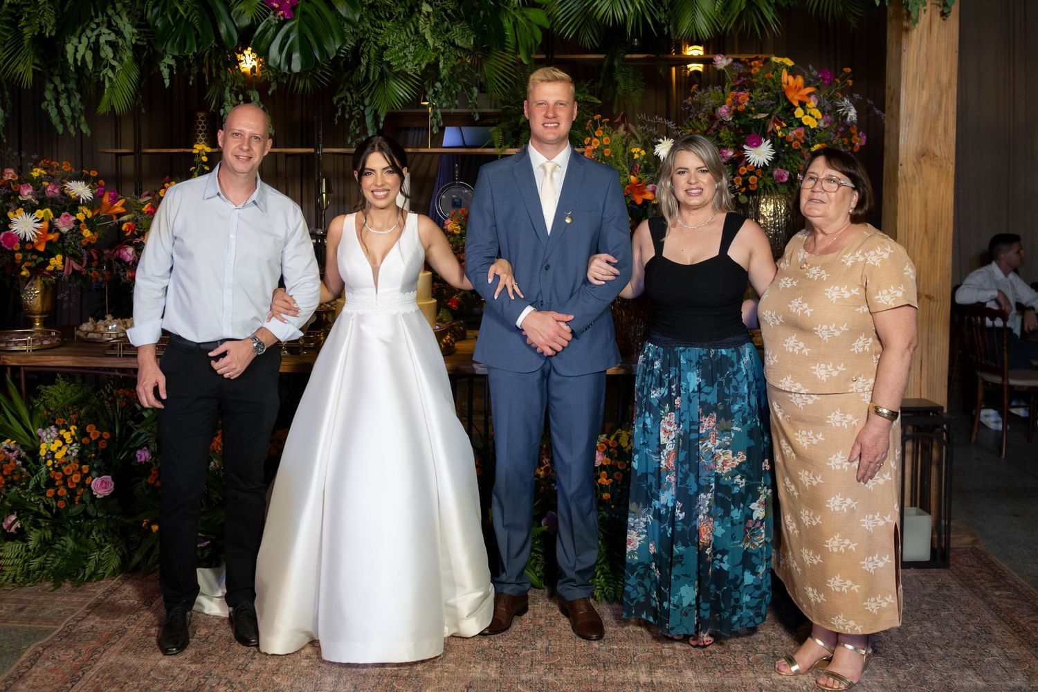 A bride in white gown and groom in blue suit pose with three guests at a floral-decorated wedding reception.