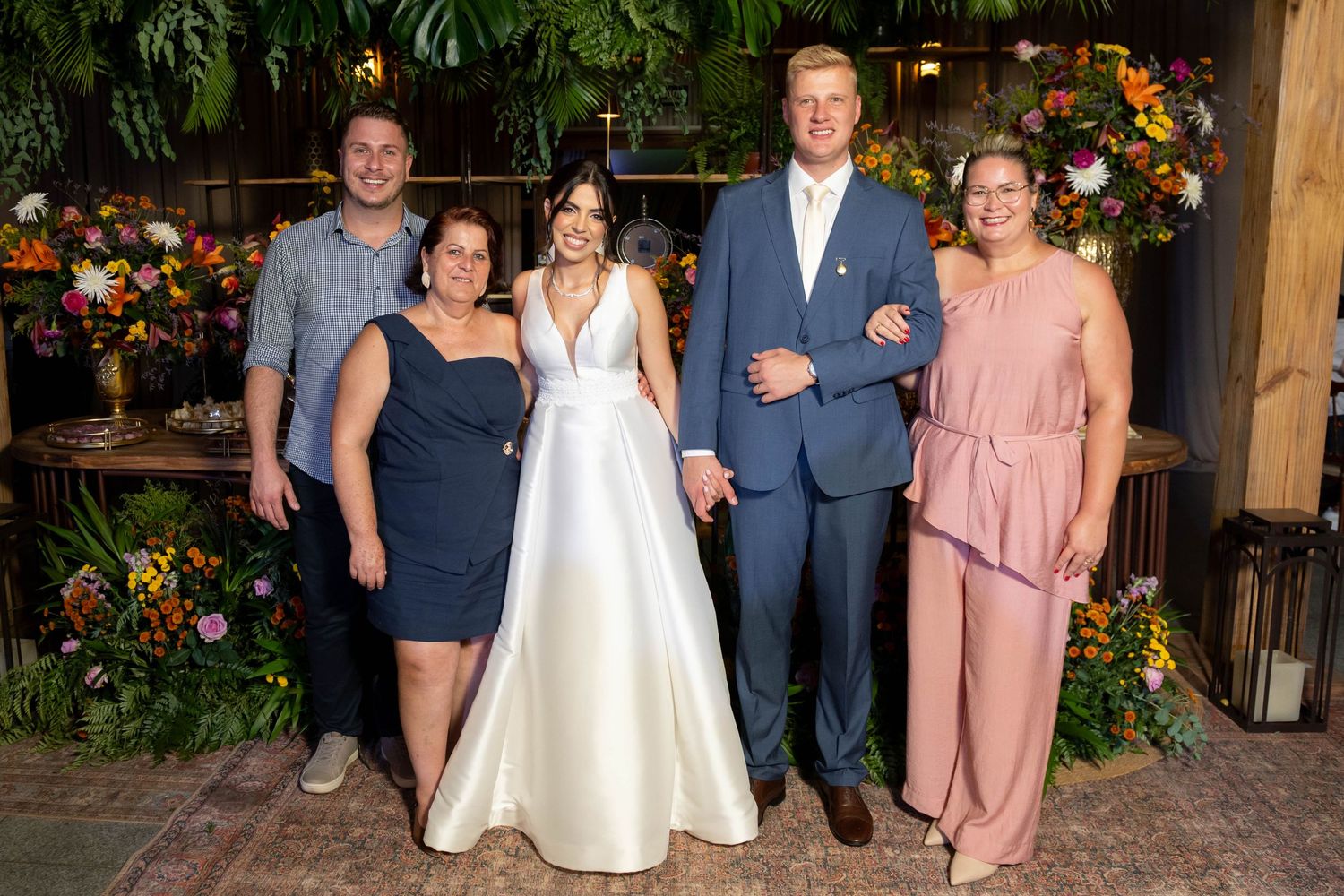 Bride and groom pose with three family members at a floral-decorated wedding reception venue.