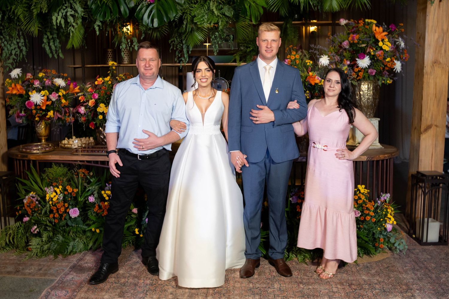Two couples pose together at a floral-decorated wedding venue, one in a white gown and blue suit, another in casual attire.