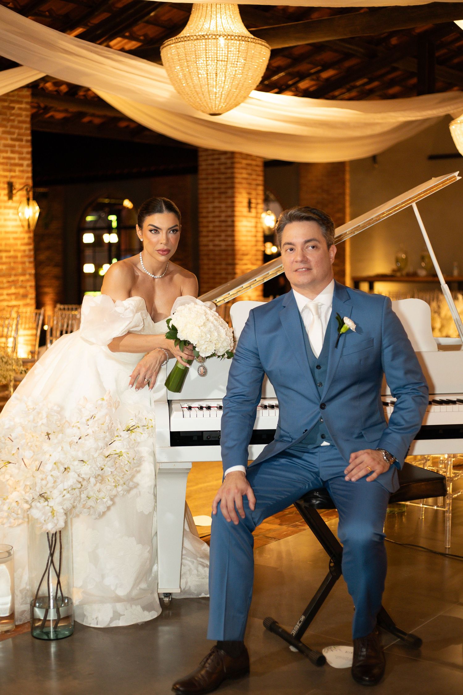 Elegant bride in white gown and groom in blue suit pose by a white piano in a warmly lit wedding venue.