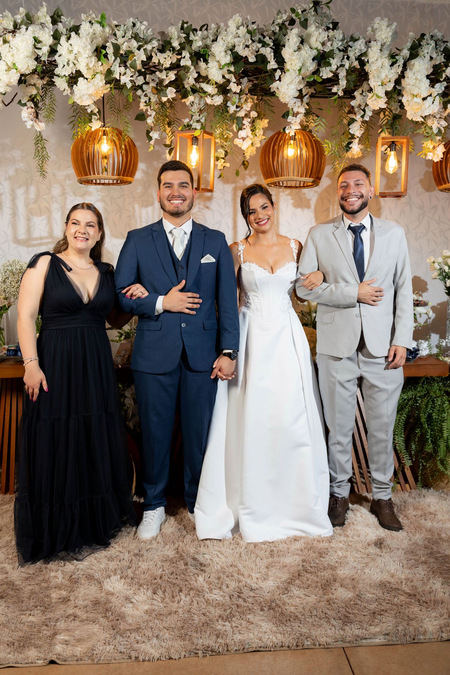 Wedding party of four posing elegantly under floral arch with warm pendant lights, bride in white gown with groom and guests.