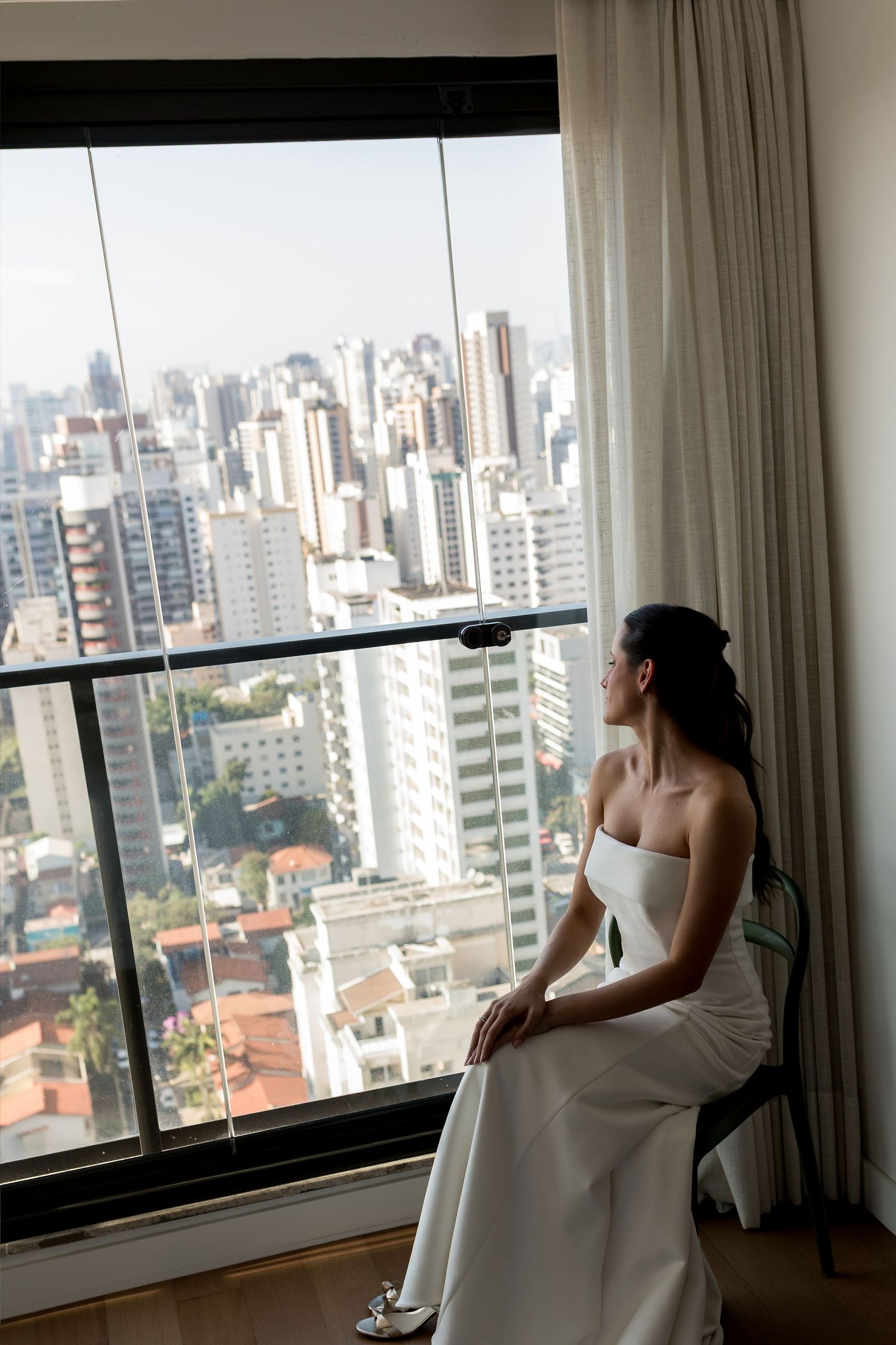 Bride in white strapless gown sits by large window overlooking city skyline on wedding day.