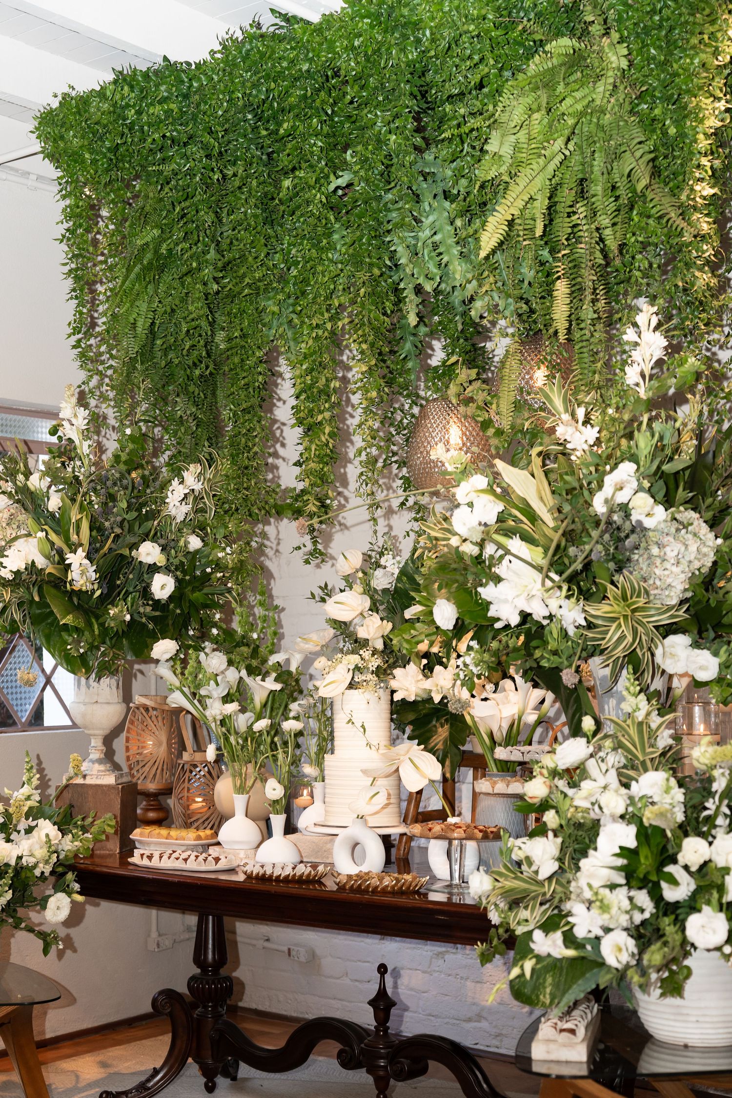 Elegant wedding dessert table with white tiered cake, surrounded by lush greenery and white floral arrangements.