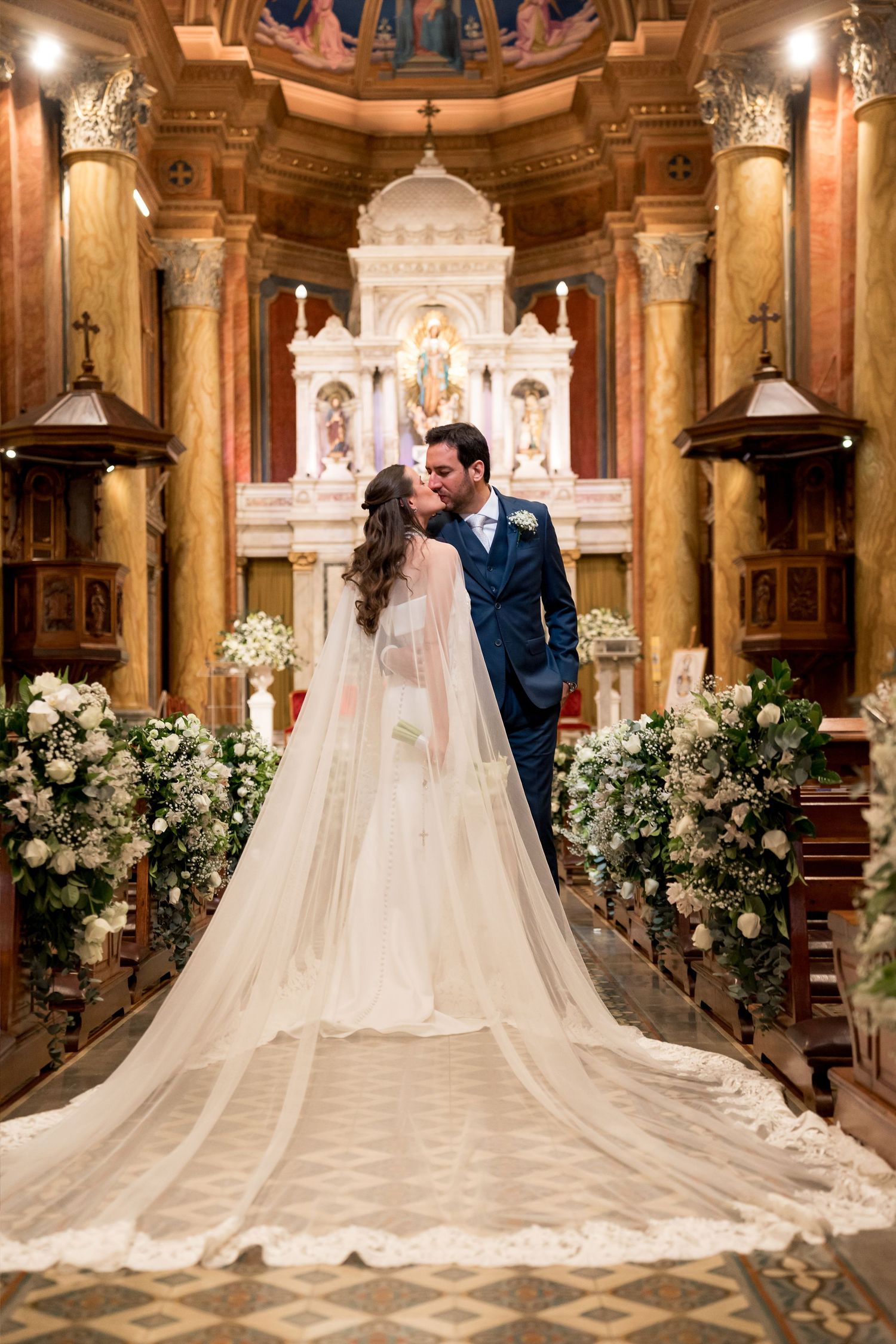 Bride and groom share first kiss at ornate church altar, surrounded by white floral arrangements, bride in cathedral veil.