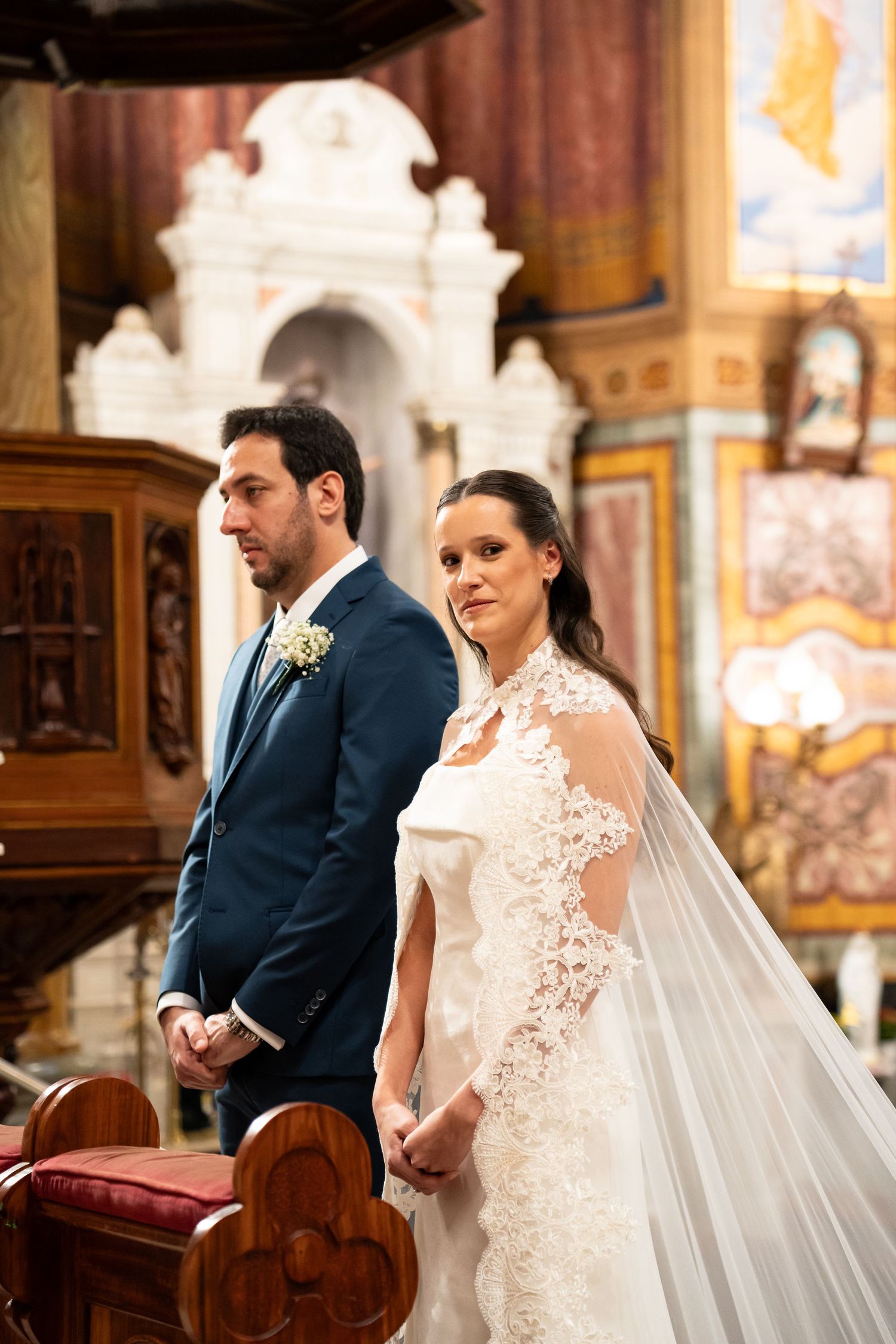 Bride in lace wedding gown with veil and groom in blue suit standing at church altar during ceremony.