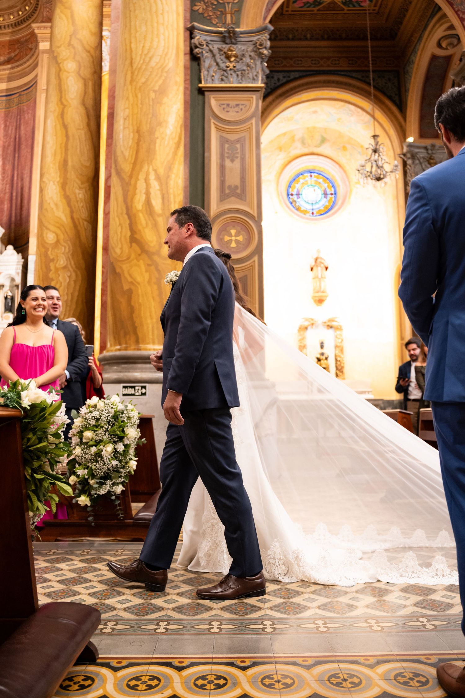 Groom in navy suit walks bride in white lace gown down church aisle during elegant wedding ceremony.