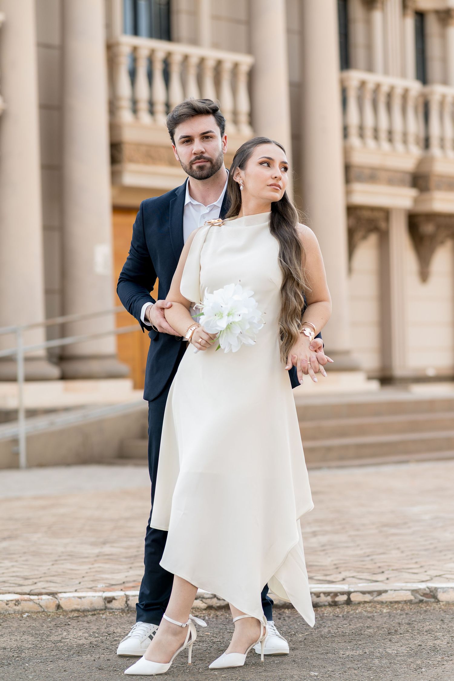 Elegant couple in wedding attire pose outside historic building, bride in white dress holding bouquet, groom in navy suit.