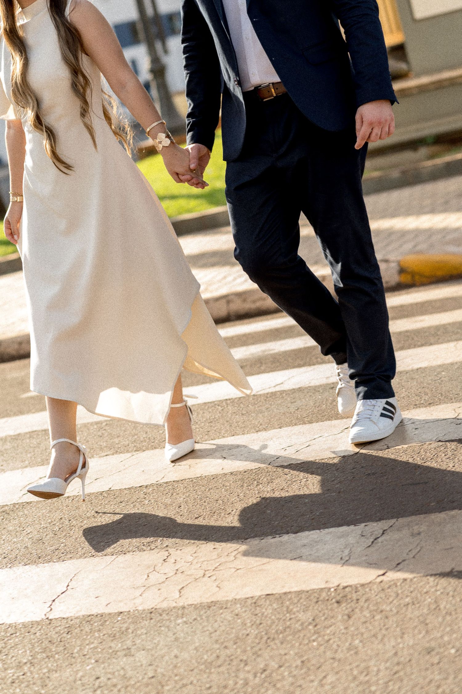 Bride and groom holding hands crossing a street crosswalk, wearing white dress and navy suit.