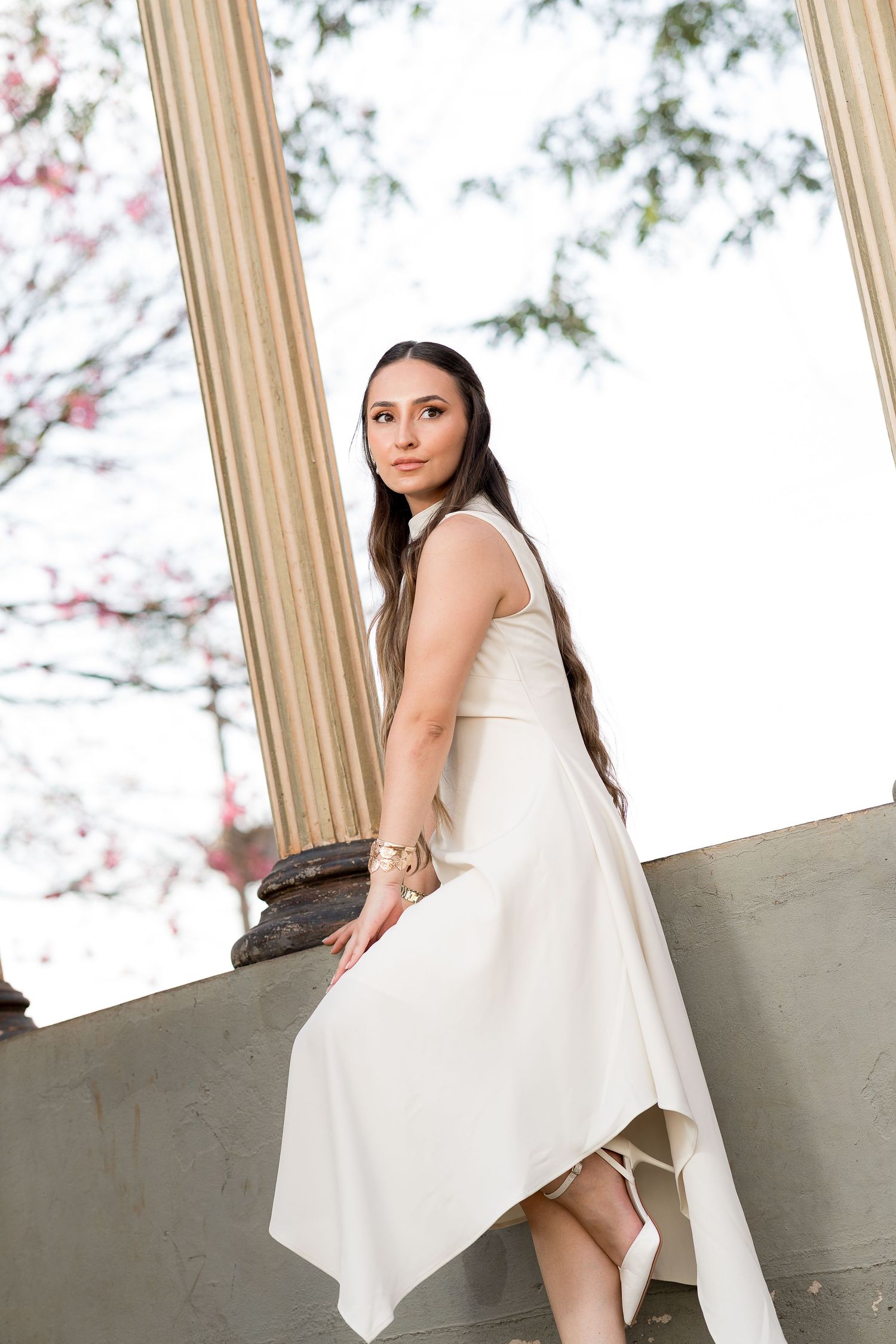 Young woman in white asymmetric dress poses elegantly by classical columns with blooming trees in background.