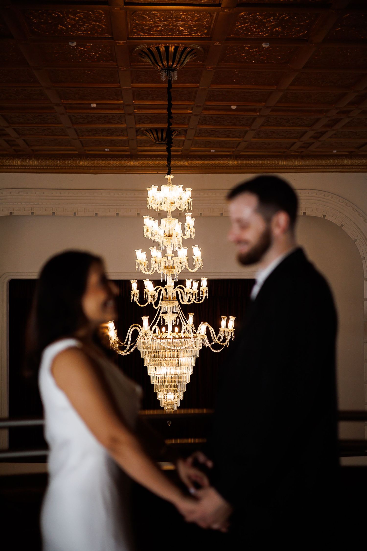 Couple holding hands in elegant venue with ornate crystal chandelier and decorative tin ceiling in background.