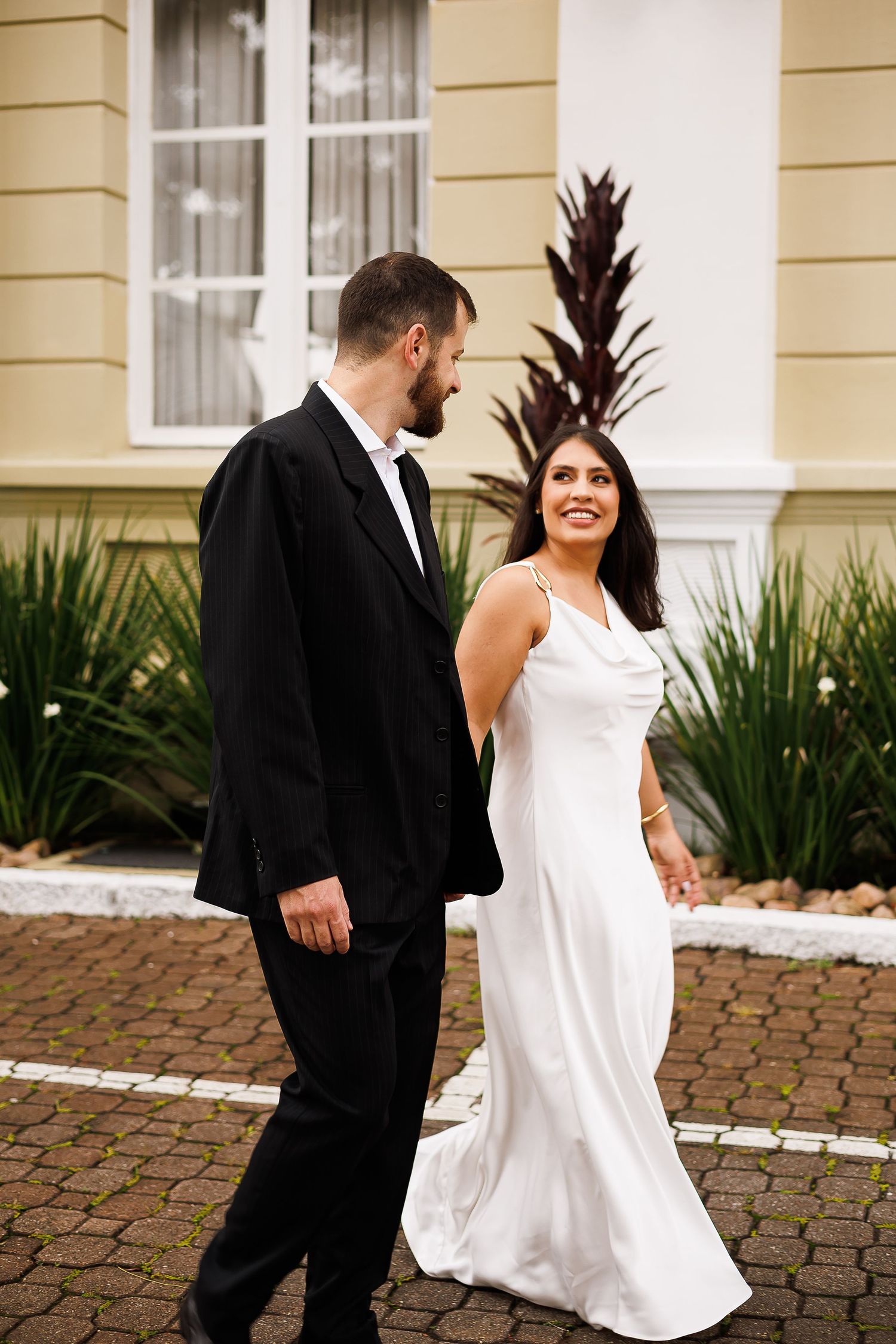 A smiling bride in a white satin gown walks with a groom in a black tuxedo outside an elegant building.