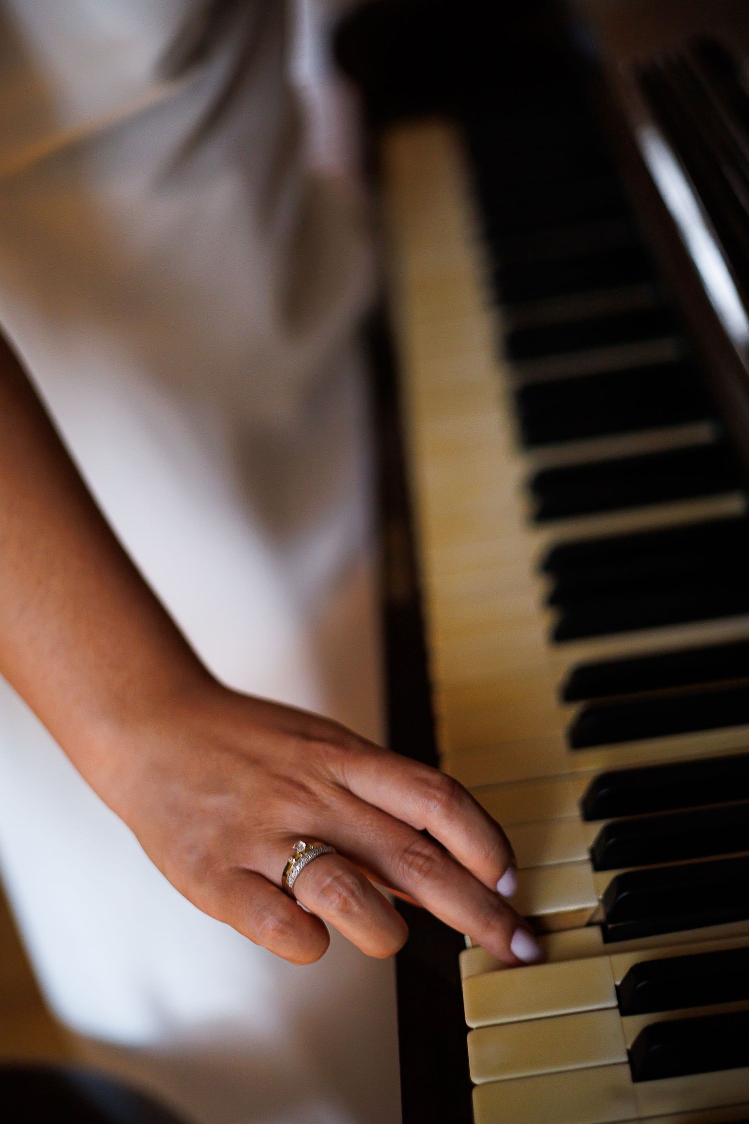 A bride's hand with engagement ring resting on piano keys, wearing a white dress.